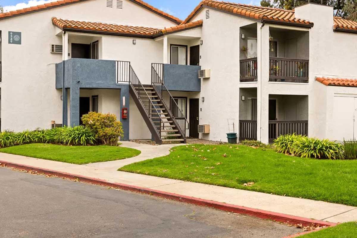 A two-story apartment building with a staircase leading to the second floor. The exterior features a light-colored stucco finish, blue accents, and a tiled roof. Lush green lawn surrounds the building, and there are small shrubs in the landscape. A pathway leads up to the entrance.