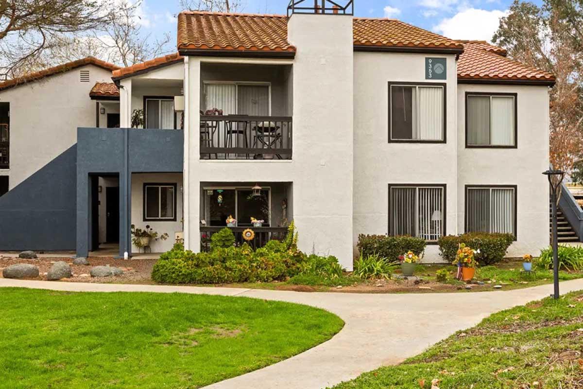 Two-story apartment building with a white exterior and brown tiled roof. The front features a small porch with black railings, surrounded by greenery and flower beds. A curved path leads to the entrance, with a grassy area in the foreground and a set of stairs leading to the upper level on the left.