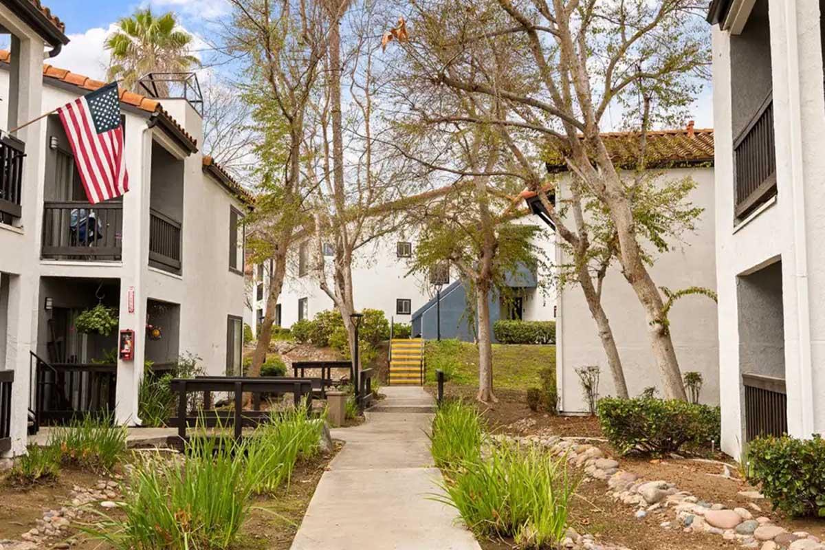 A pathway leads through a residential area with two-story buildings featuring balconies. American flags hang from one of the balconies. There are trees and shrubs lining the walkway, with a staircase visible in the background, creating a peaceful atmosphere in the community.