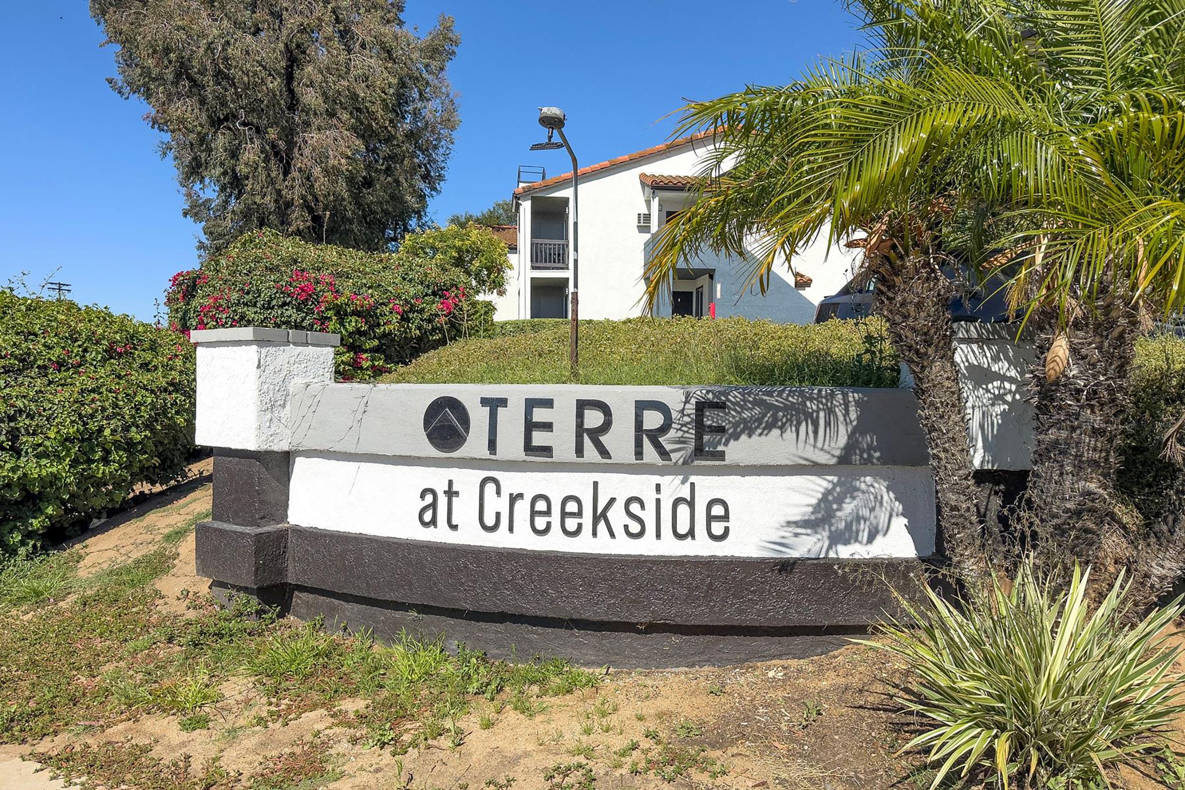 Sign for "Terre at Creekside" located near a landscaped area with greenery and palm trees, alongside a light fixture. A multi-story building can be seen in the background under a clear blue sky.