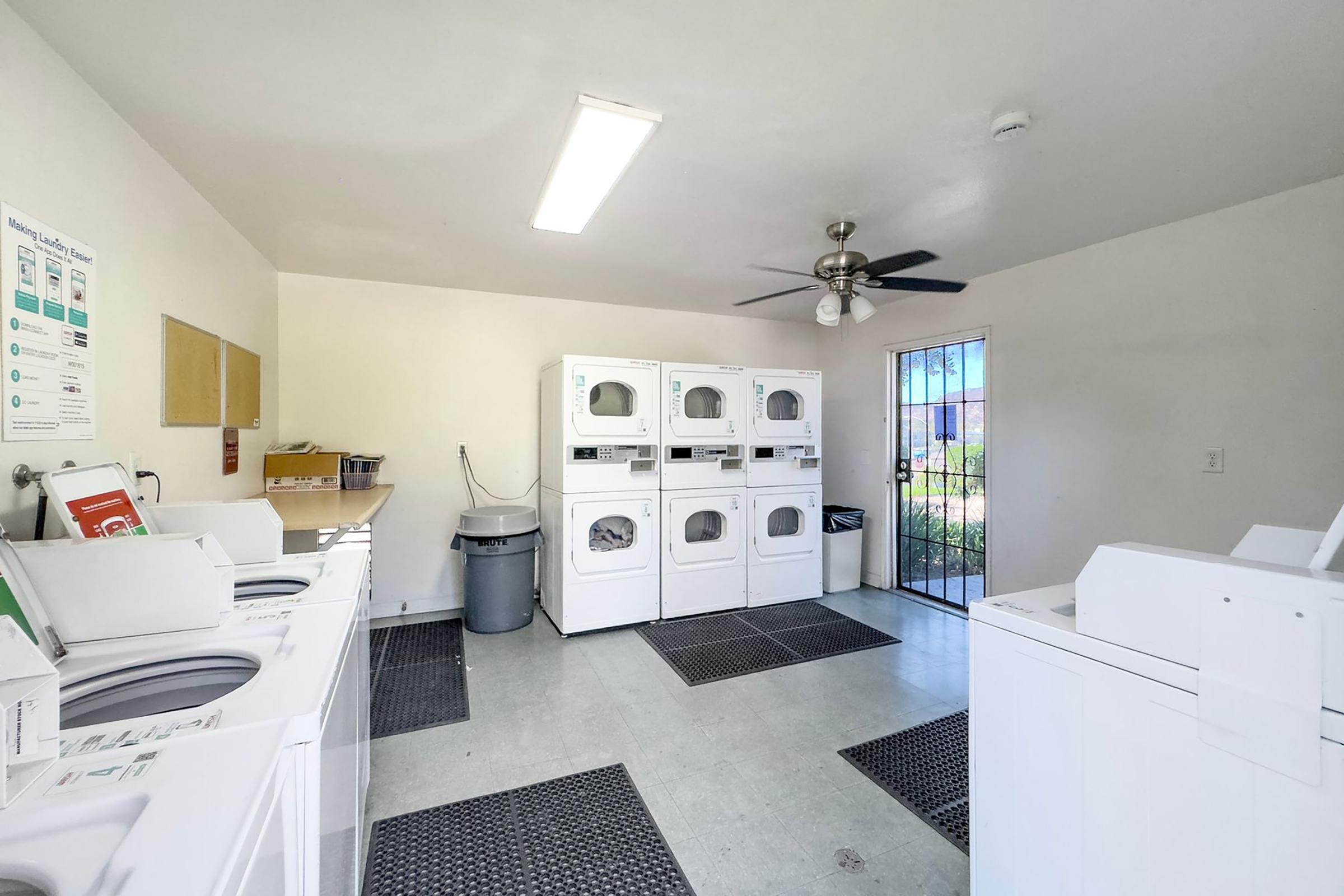 Laundry room with multiple white washing machines and dryers arranged against the wall. There is a gray trash bin in the corner and a ceiling fan overhead. The room is well-lit with natural light coming through a windowed door leading outside. Floor mats are placed in front of the machines.