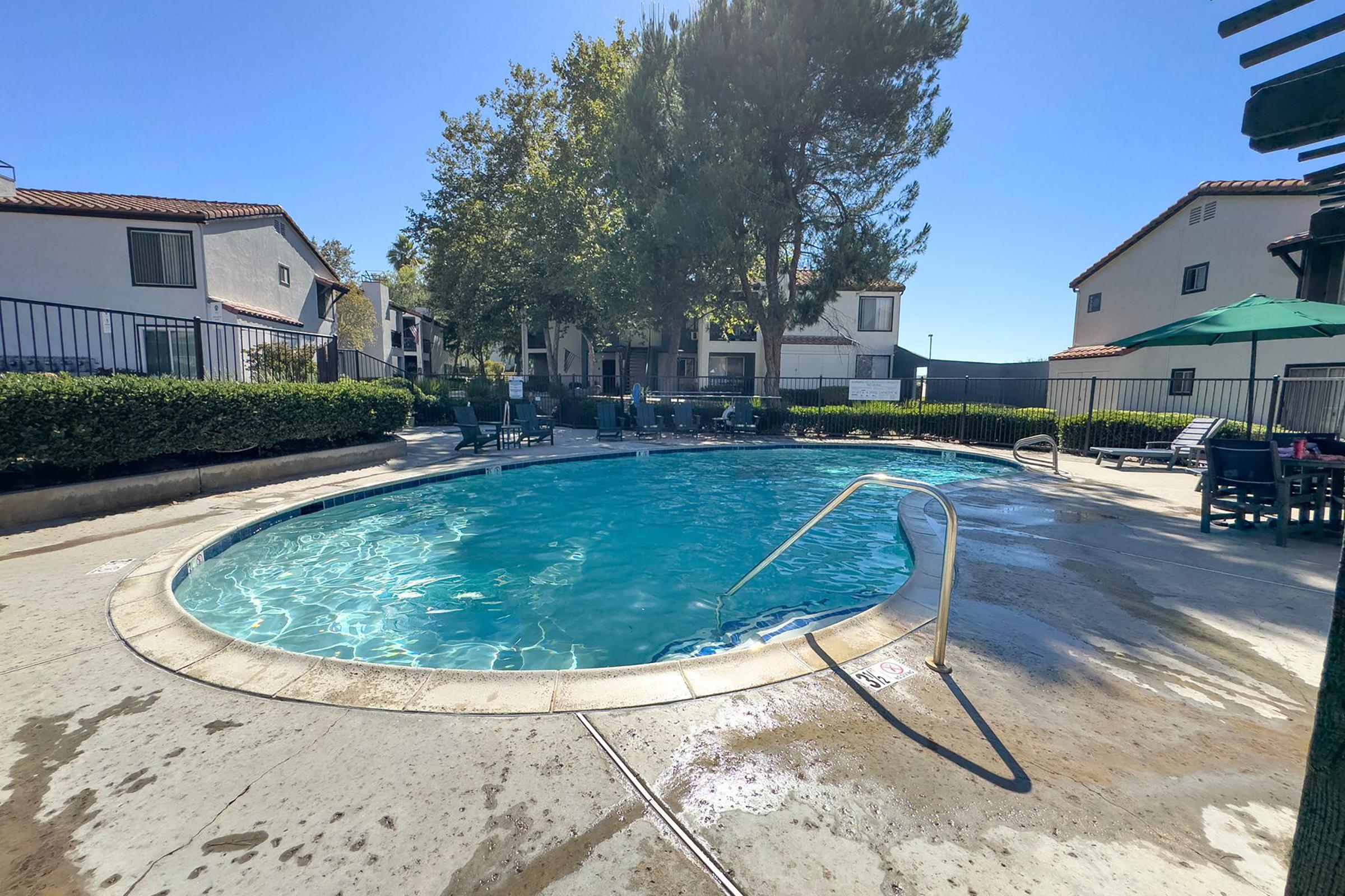 A swimming pool surrounded by greenery and lounge chairs, with a clear blue sky overhead. The pool has a curved shape and features a ladder for entry. Apartment buildings are seen in the background, and there are umbrellas for shade. The scene conveys a relaxing outdoor atmosphere.