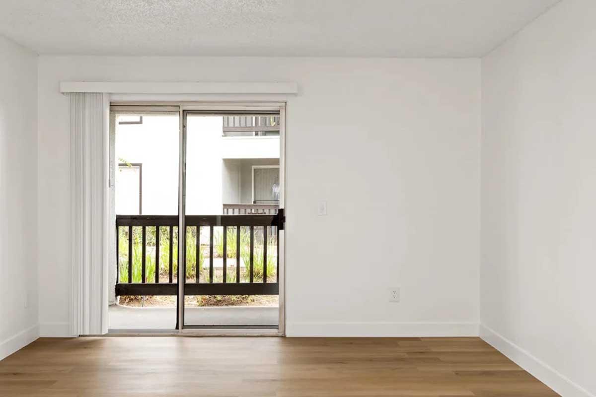 A spacious, empty room with white walls and a wooden floor. Large glass sliding doors lead to a balcony area with greenery visible outside. Natural light fills the space, creating a bright and open atmosphere.