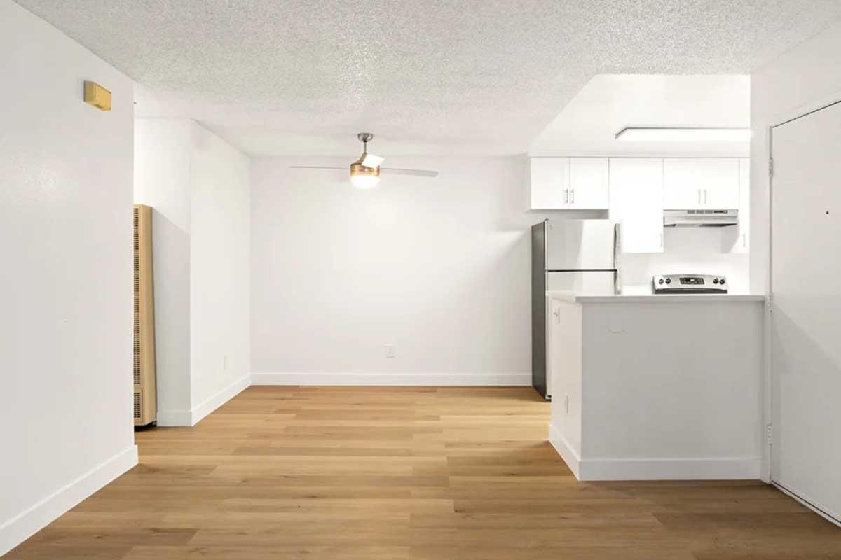 A bright and modern living space featuring light wood flooring, a ceiling fan, and a kitchen area with white cabinetry, stainless steel appliances, and a counter. The walls are painted white, creating an airy atmosphere. The room is empty, ready for personal touches.