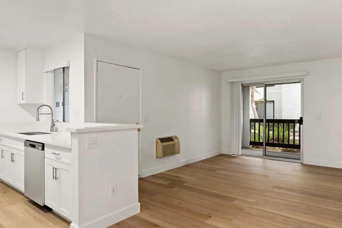 Interior view of a modern, bright living space featuring a kitchen area with stainless steel appliances, a door leading outside, and large windows. The flooring is light wood, and the walls are painted white, creating an open, airy atmosphere.