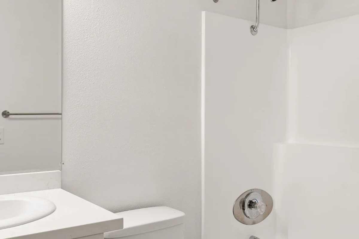 A clean, minimalistic bathroom featuring a white shower stall, a toilet, and a sink with a mirror above it. The walls are plain and light-colored, and there is a towel bar mounted on the wall. The overall aesthetic is simple and contemporary.