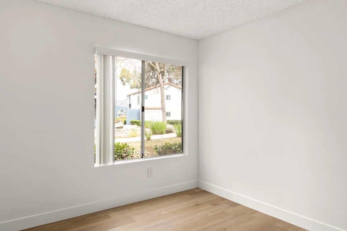 A bright, empty room with light-colored walls and wooden flooring. A large window provides a view of a garden and houses outside, allowing natural light to fill the space. The room has simple decor, emphasizing a modern and minimalist aesthetic.