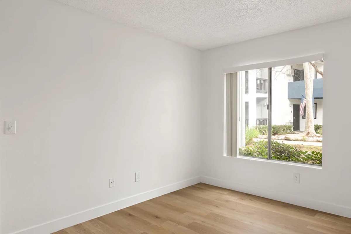 A bright, empty room with white walls and a large window featuring a view of greenery outside. The floor is made of light-colored wood, and the window has a simple roller blind partially open, allowing natural light to fill the space.