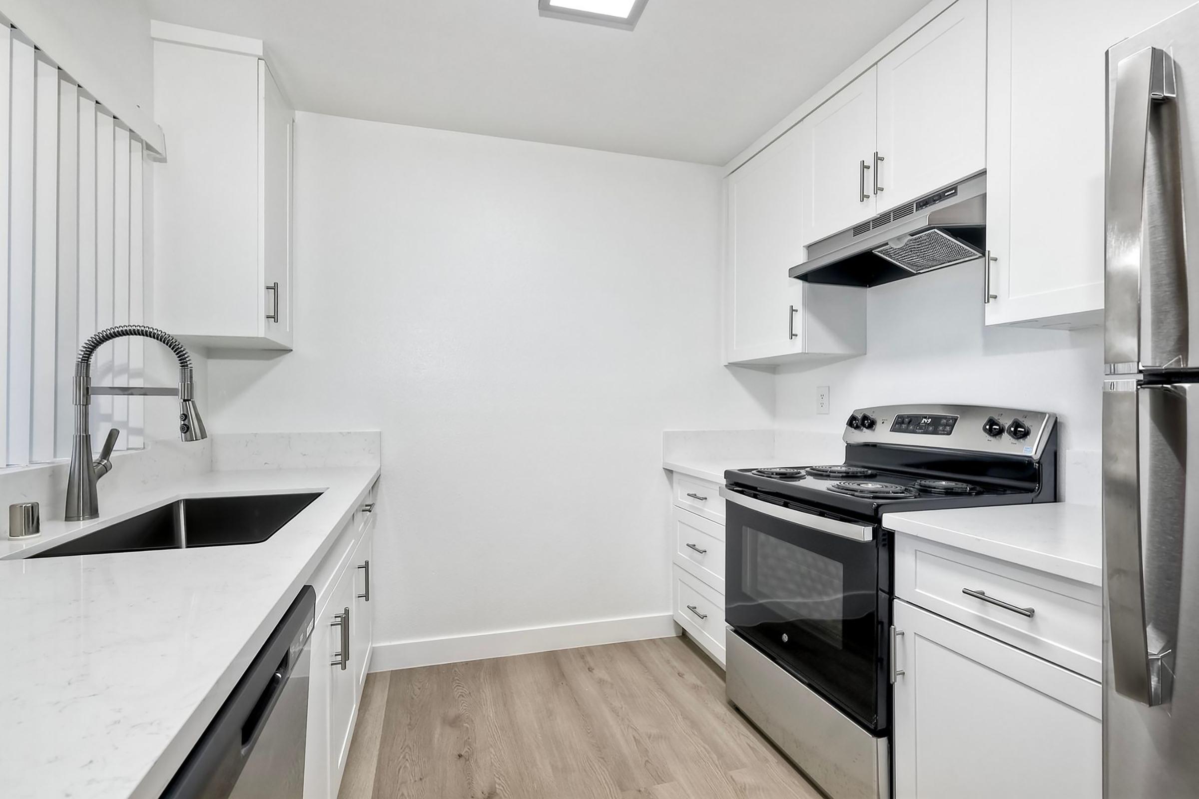 A modern kitchen featuring white cabinetry, a stainless-steel refrigerator, a black electric stove with a hood, a double-basin sink, and light-colored countertops. The floor is wooden and the walls are white, creating a bright and clean atmosphere.