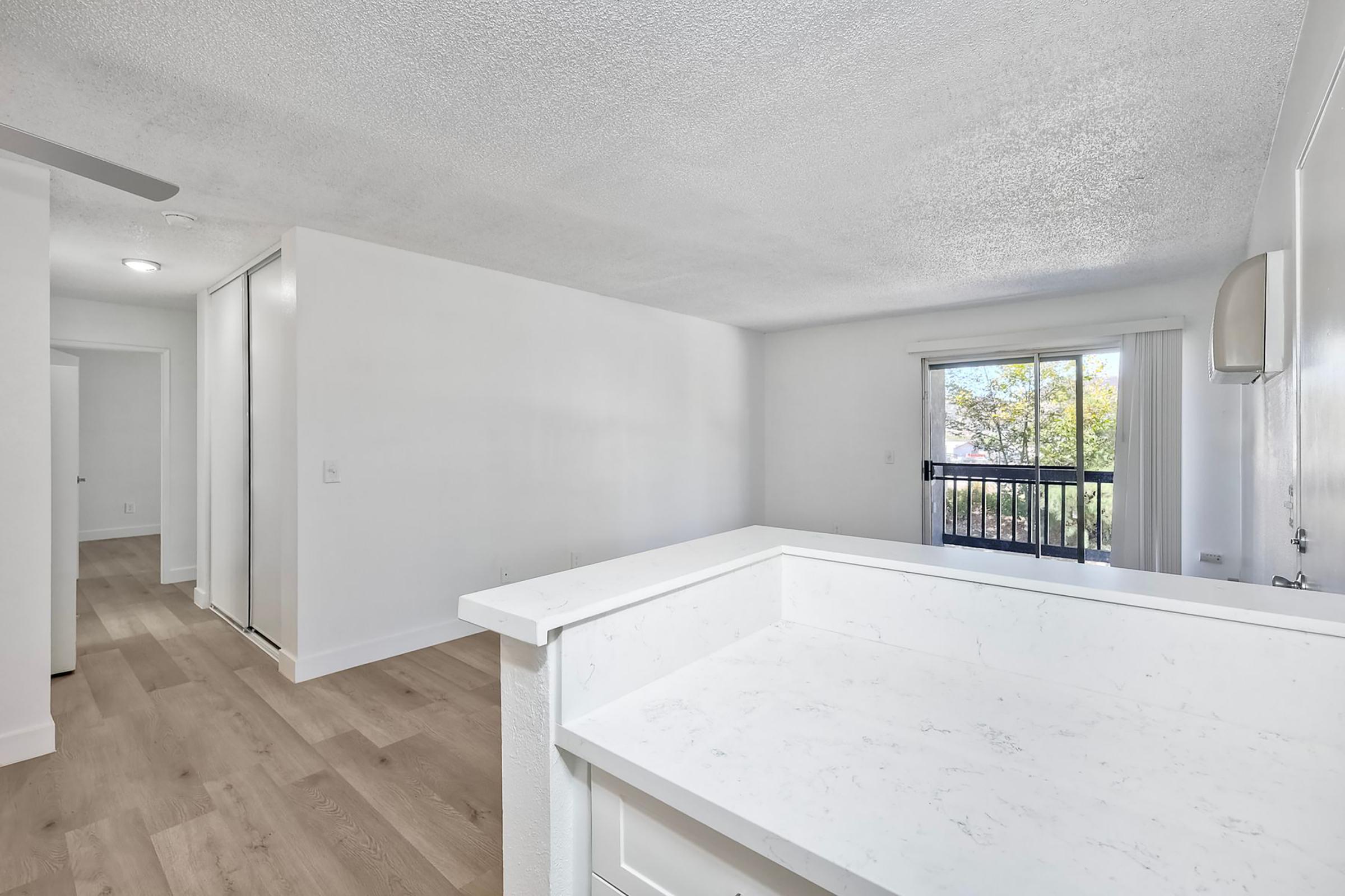 Bright and airy interior of a modern living space. Features a white kitchen counter overlooking a spacious living area with light wood flooring, natural light from a window, and a balcony view. Simple and clean design with neutral colors.