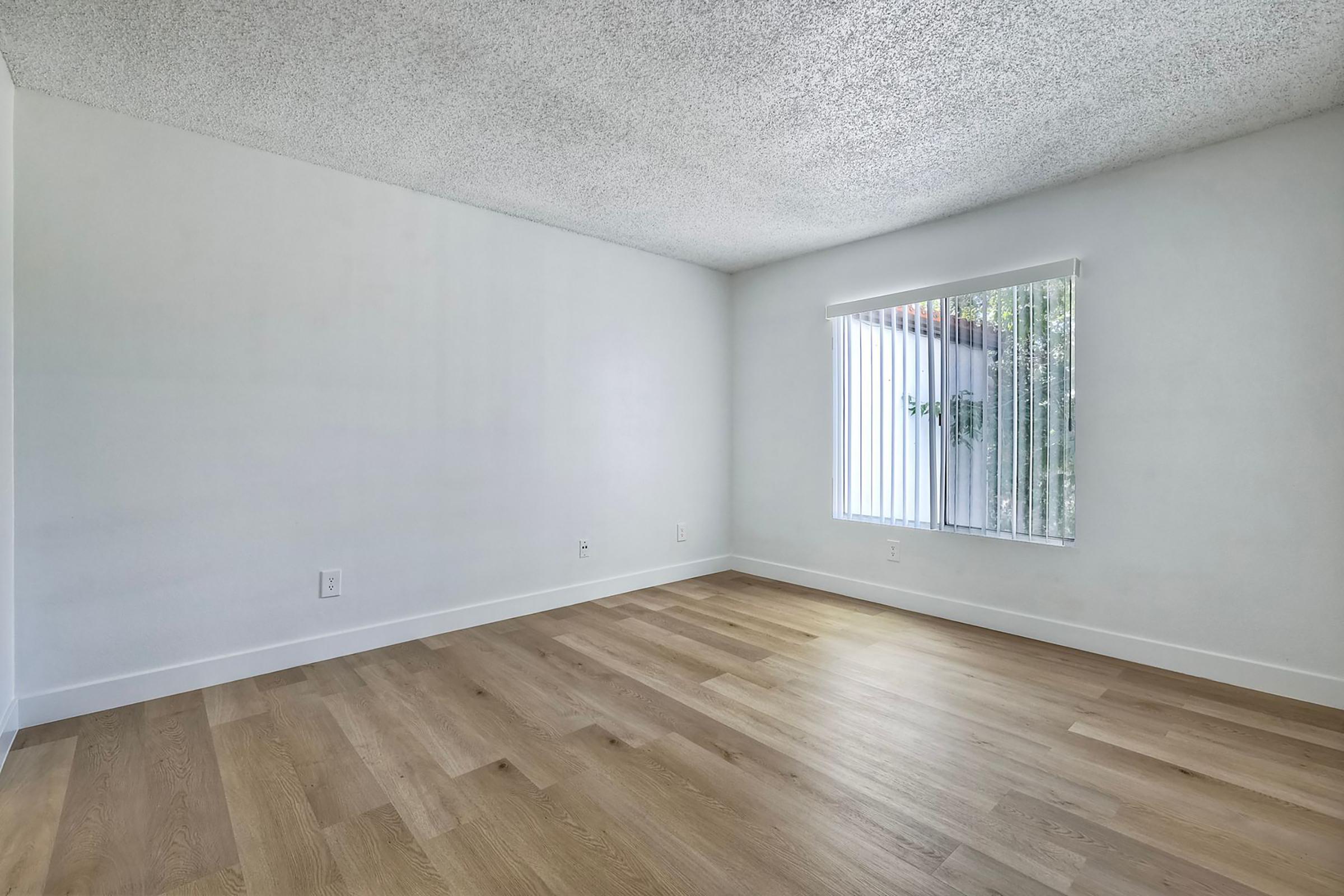 Empty room with light-colored walls and a textured ceiling. The floor is covered with wood-like laminate. A window with vertical blinds allows natural light to enter, revealing a view of greenery outside. The space appears clean and ready for furnishing.