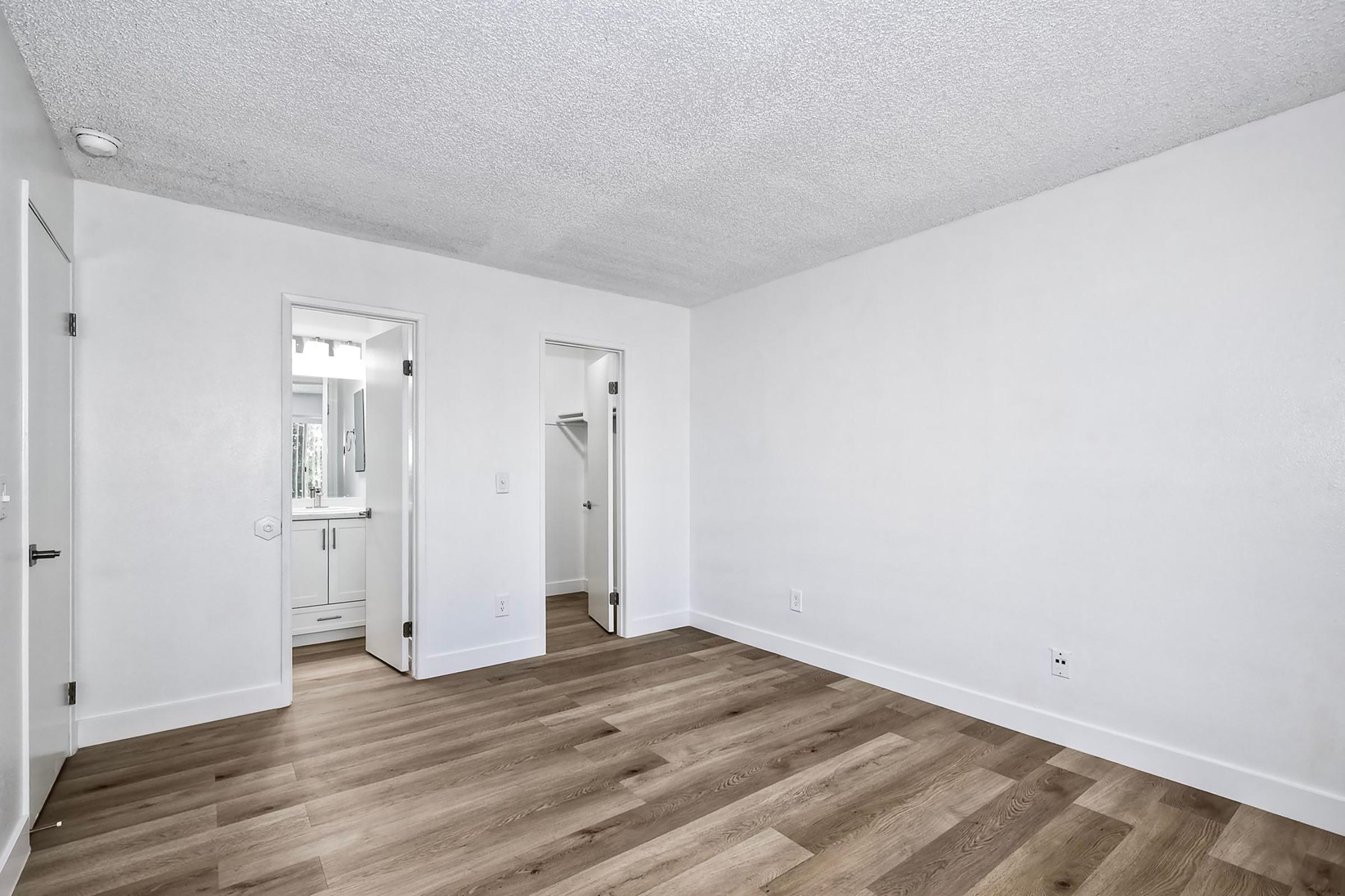 A spacious, empty room with light-colored walls and hardwood flooring. Two doorways lead to adjoining areas, one of which is a bathroom with a window. The room has a minimalist design, with no furniture or decorations, providing a neutral and airy atmosphere.
