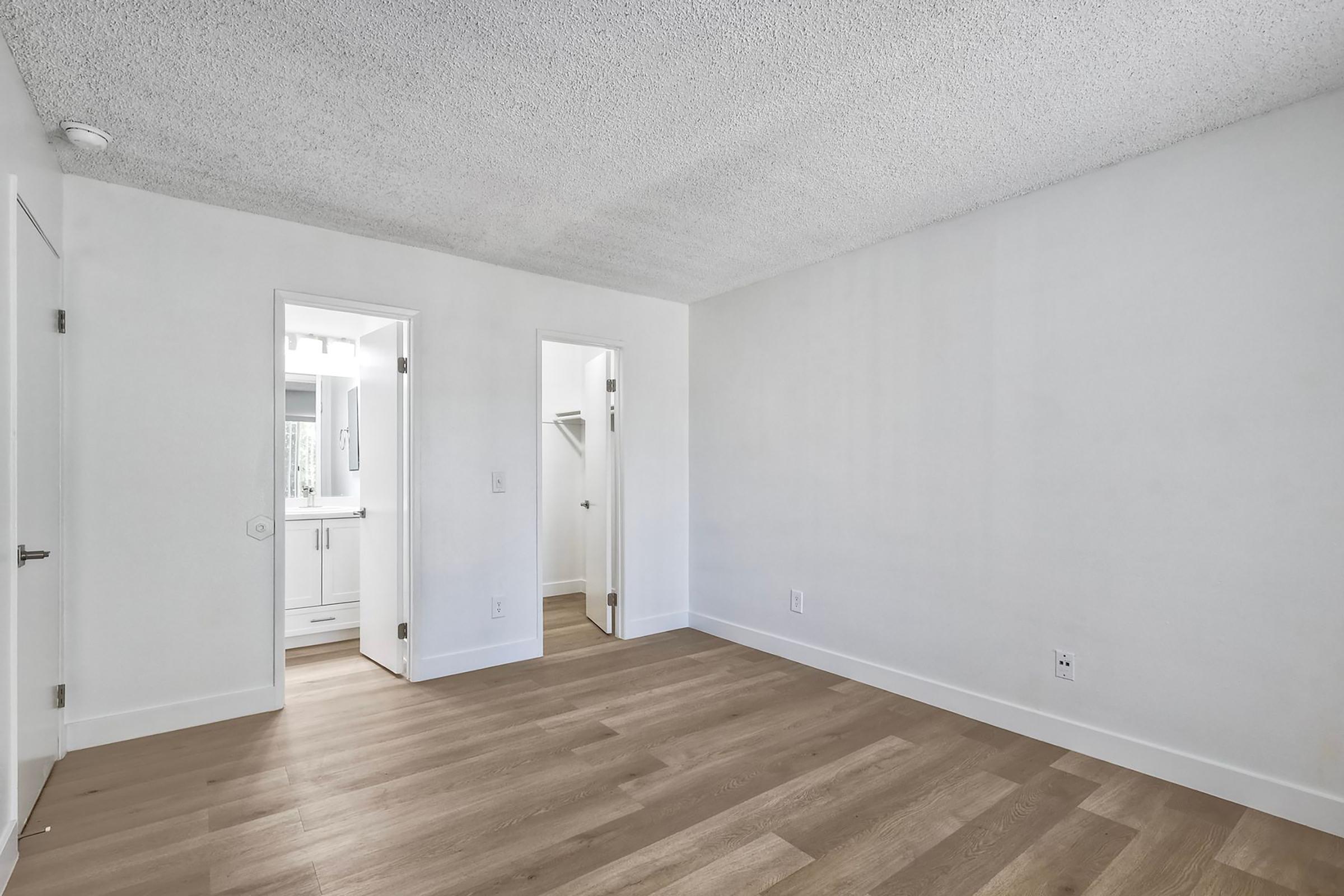 A spacious, empty room with light gray walls and a textured ceiling, featuring wood laminate flooring. Two doorways are visible, leading to a bathroom and a closet. The room is well-lit, with natural light coming from the adjacent area.