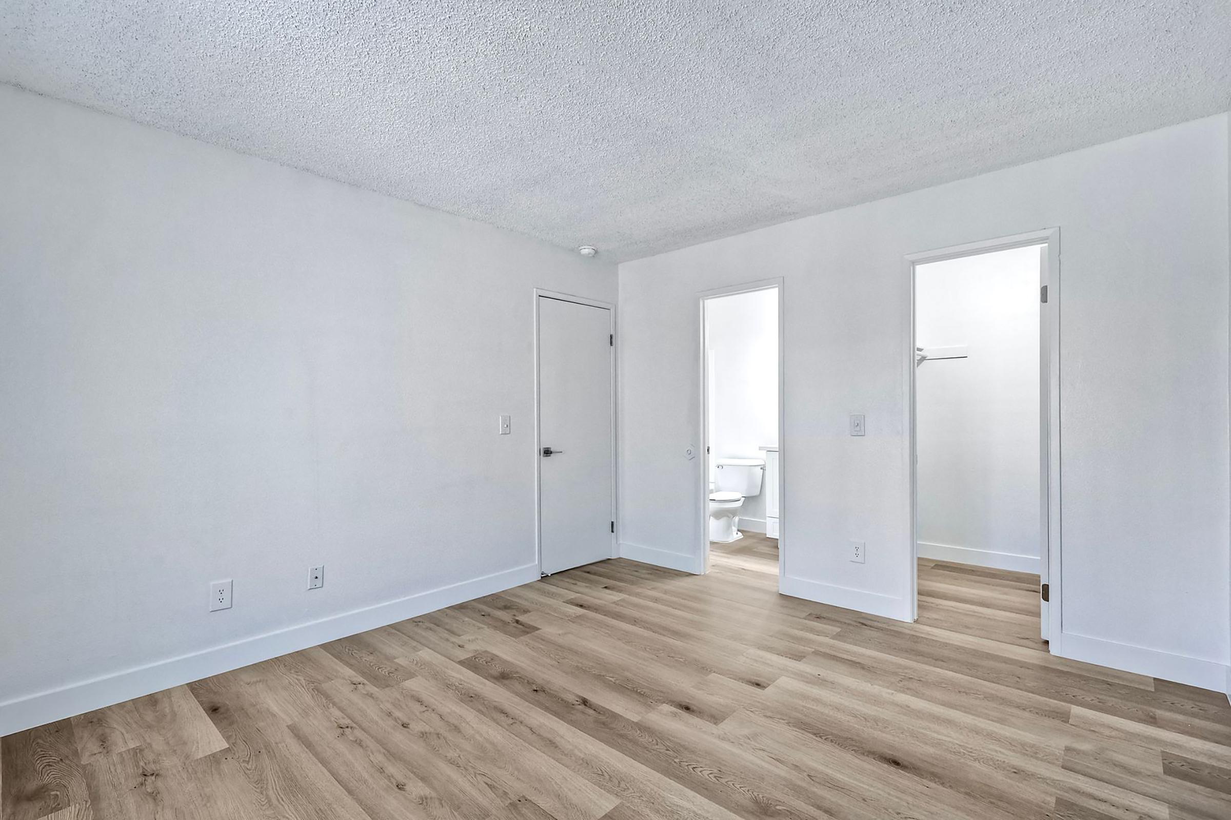 Empty room with light-colored walls and wooden floor. There are two doorways, one leading to a bathroom and the other to another room. The ceiling is textured, and the overall ambiance is bright and spacious.