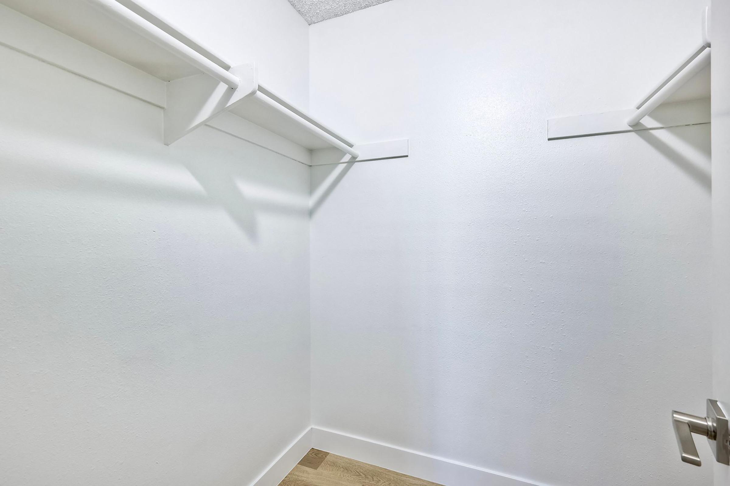 Empty closet space with white walls and two horizontal shelves. The floor is light-colored wood, and there is a door partially visible on the right side. The overall look is clean and minimalistic, suitable for storage.