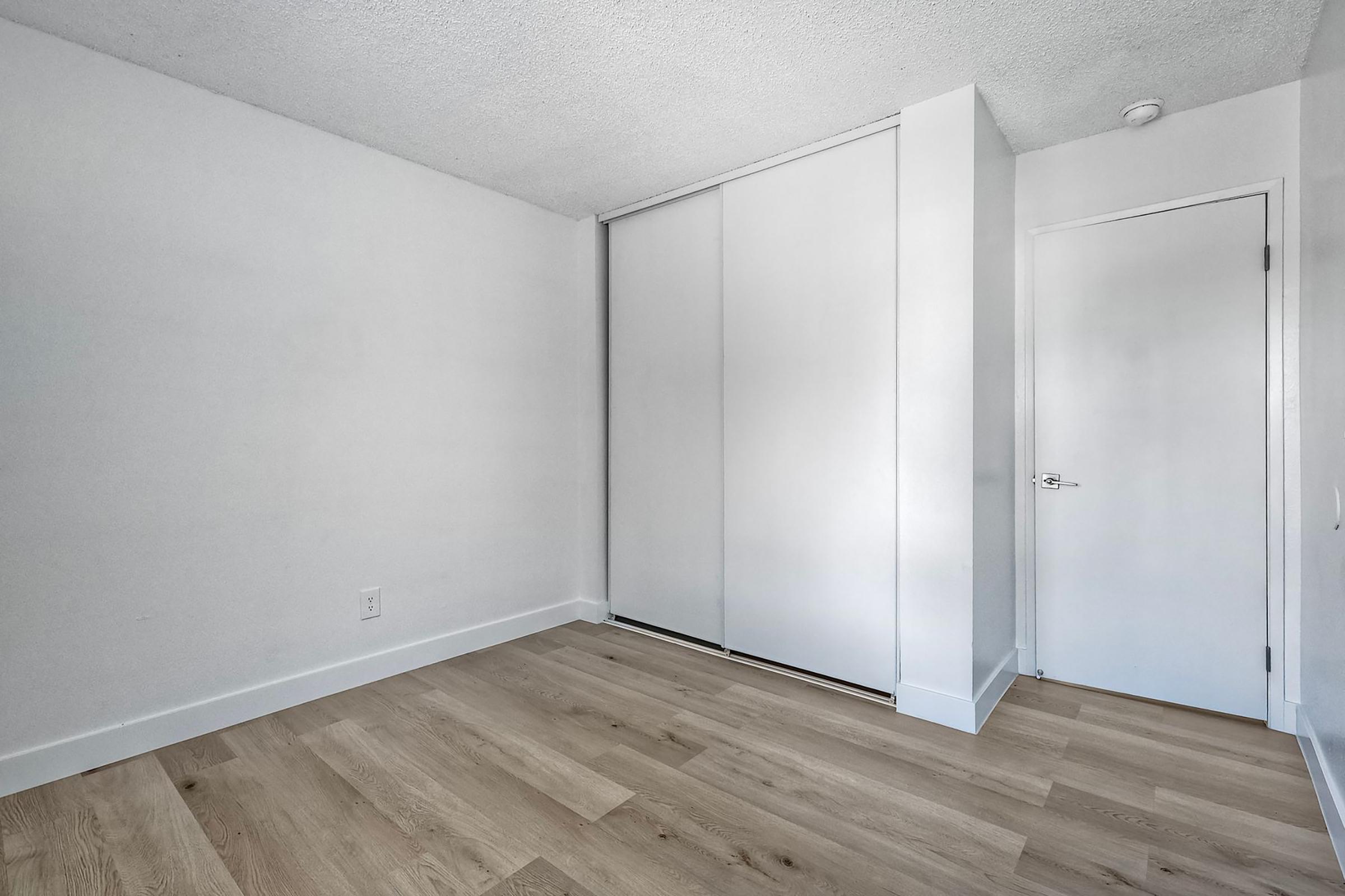 A clean, minimalistic room with light-colored walls and hardwood flooring. On the left, there are sliding closet doors, and a white door is visible on the right. The space is well-lit, creating an open and airy atmosphere.