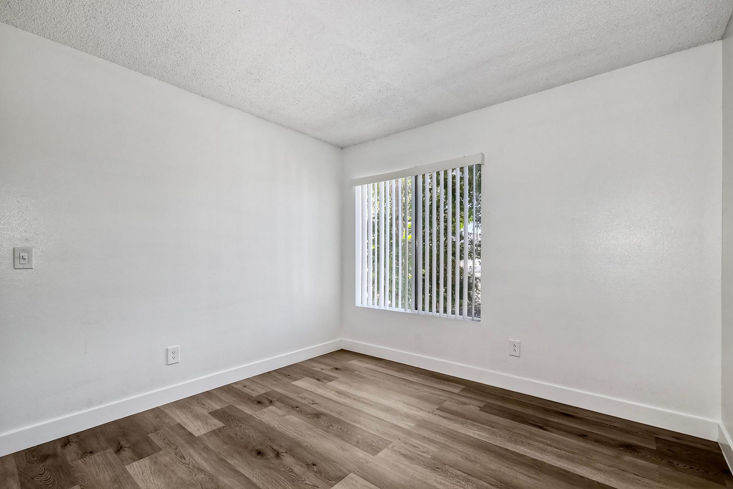 Empty room with a light-colored textured ceiling, white walls, and laminate flooring. A large window with vertical blinds allows natural light to enter, revealing greenery outside. The space is minimalist and unfurnished, emphasizing the open layout.