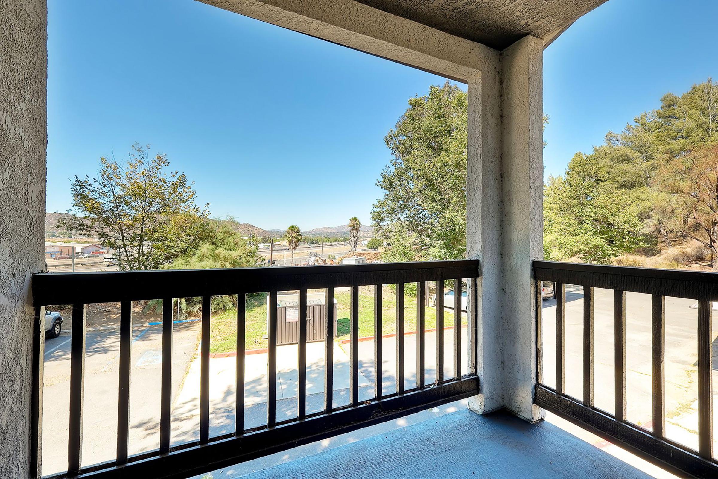 View from a balcony overlooking a serene outdoor landscape with trees, a small road, and distant hills under a clear blue sky. The railing is black, and the floor is painted blue, creating a cozy outdoor space for relaxation.