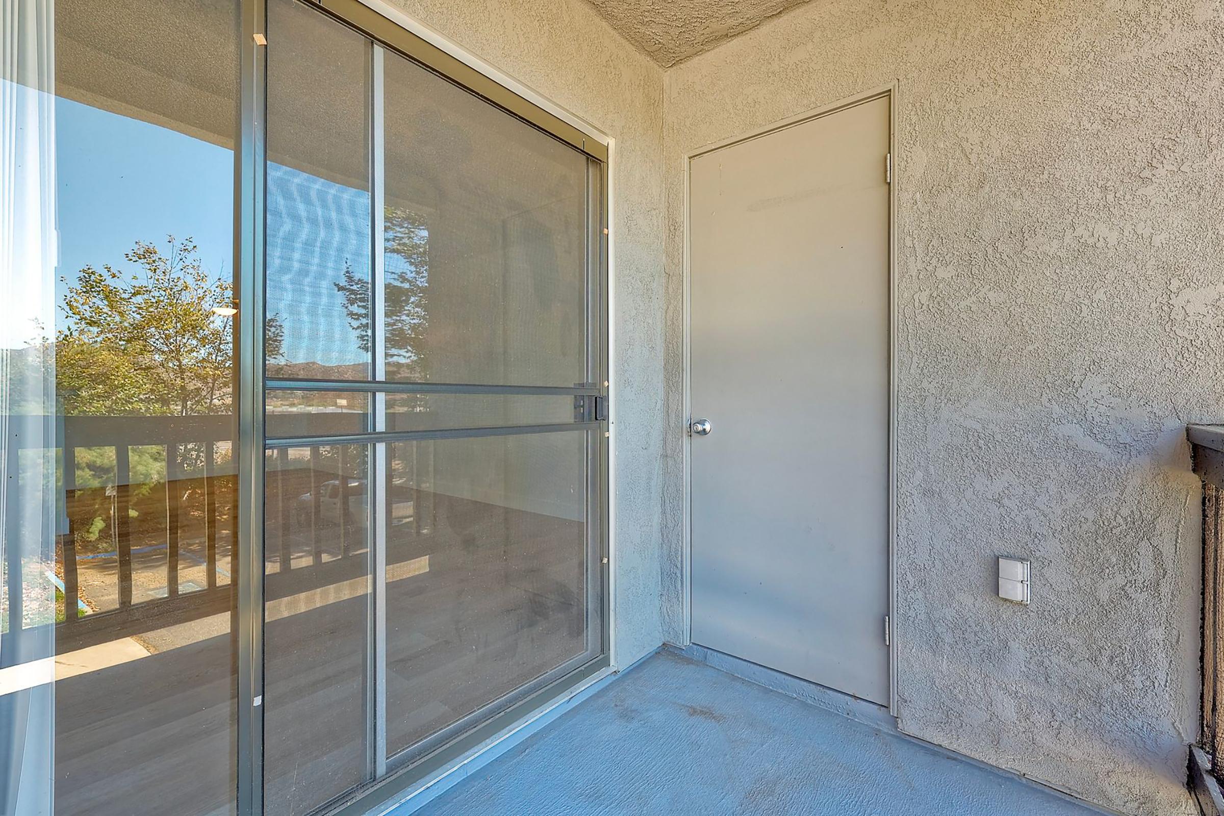 A view of a balcony featuring a sliding glass door and a closed door leading to another room. The balcony floor is grey and the walls are textured concrete. Natural light is coming through the glass door, and there are trees visible in the background.