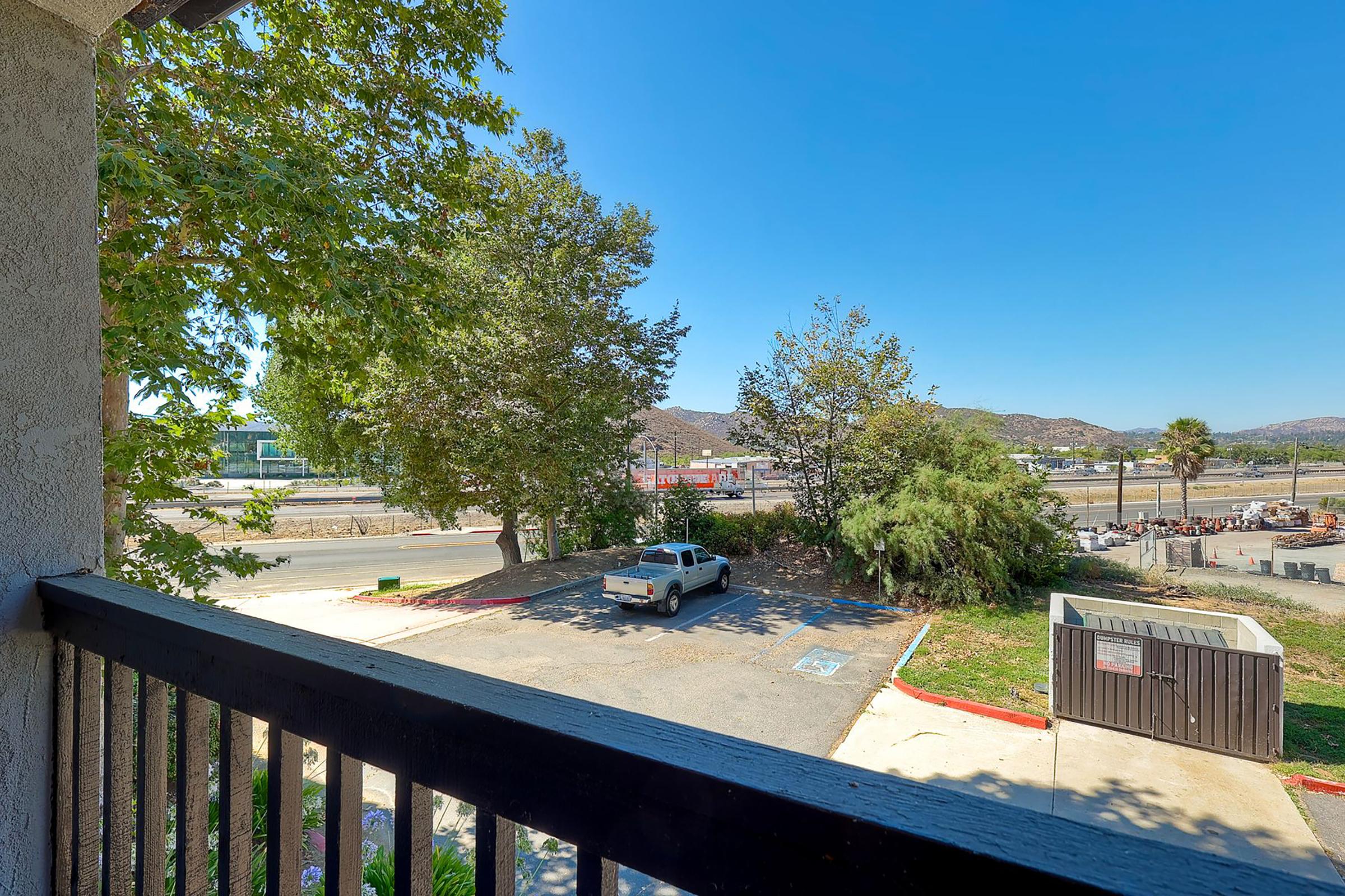 View from a balcony overlooking a parking area with a white pickup truck. In the background, there are trees, a road, and distant hills under a clear blue sky. A dumpster is visible on the right side of the image. The scene suggests an outdoor, suburban environment.