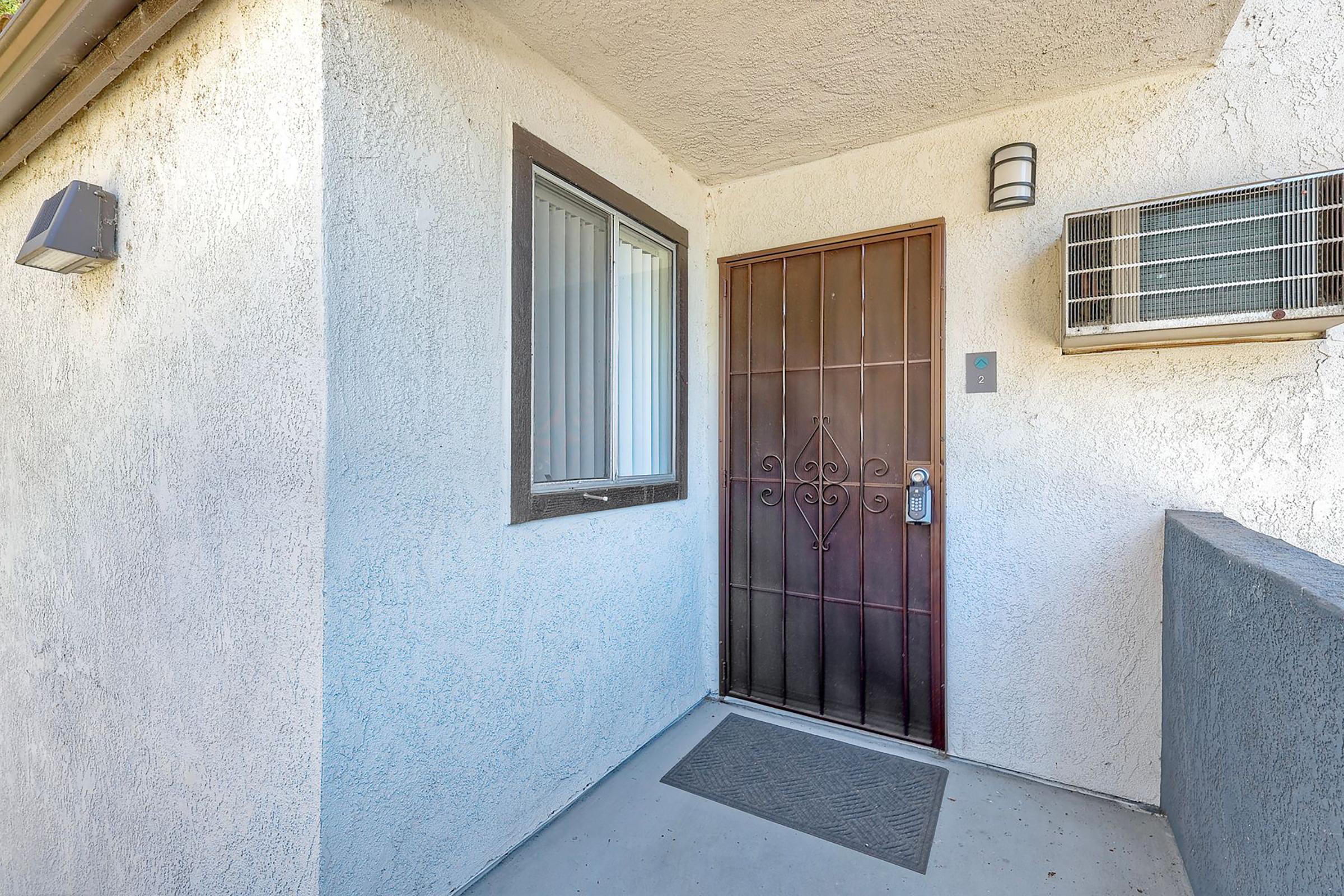 Entrance of a residential unit featuring a brown door with decorative metalwork, a small window to the left, and an air conditioning unit mounted above. The walls are painted light beige, and there is a doormat on the floor.