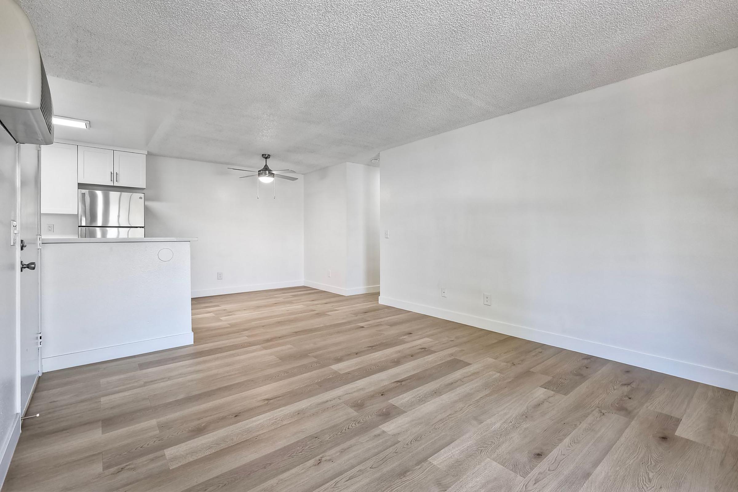 Spacious and bright interior of an apartment featuring light wood flooring, white walls, and a modern kitchen area with stainless steel appliances. A ceiling fan is visible, and the room is well-lit, creating an open and inviting living space.