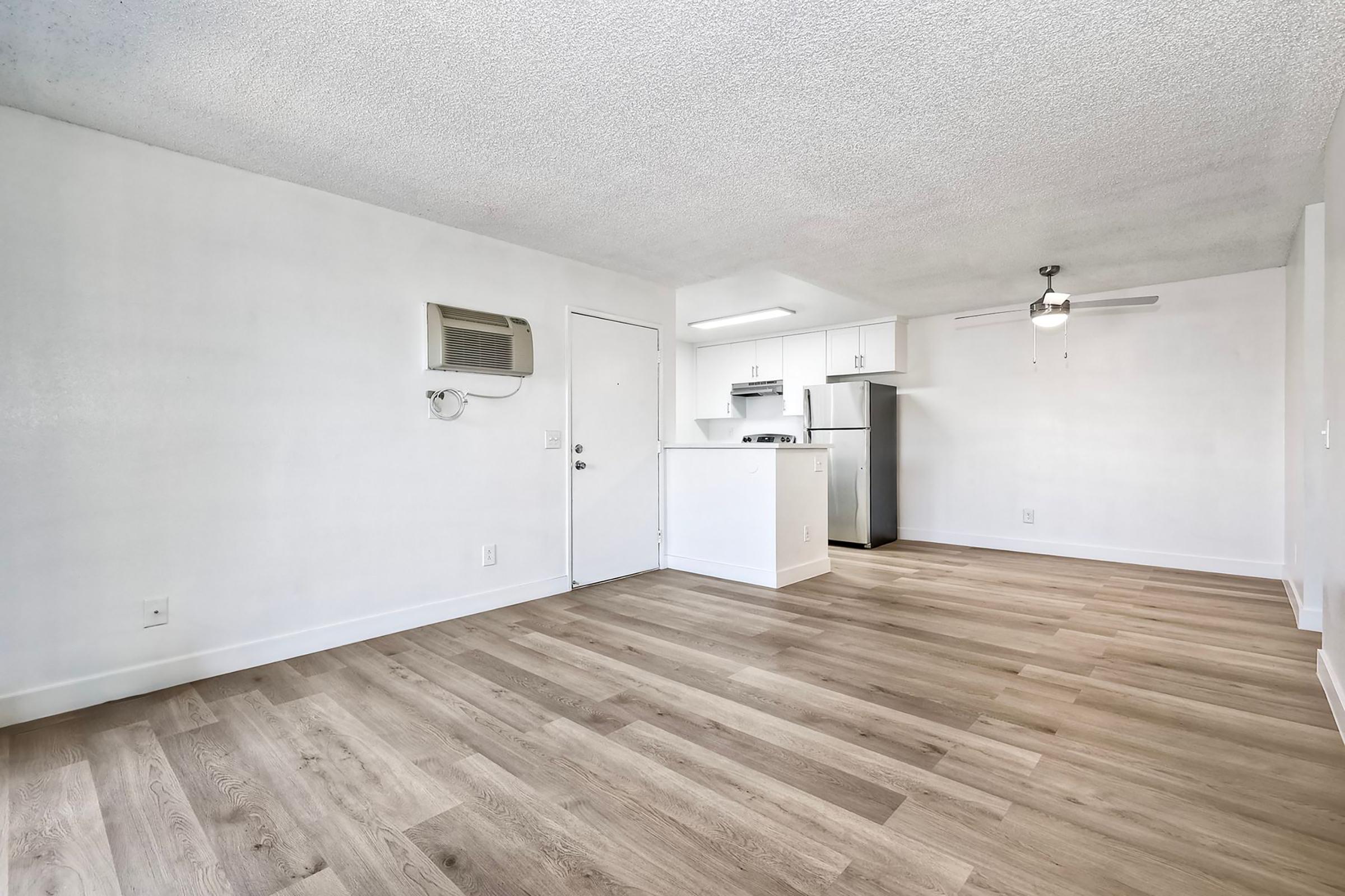 Bright, spacious interior of a modern apartment. The room features light-colored walls and wooden flooring. An open kitchen area with white cabinetry and stainless steel appliances is visible. A ceiling fan is mounted in the corner, and there's a door leading to the entrance. Natural light fills the space.