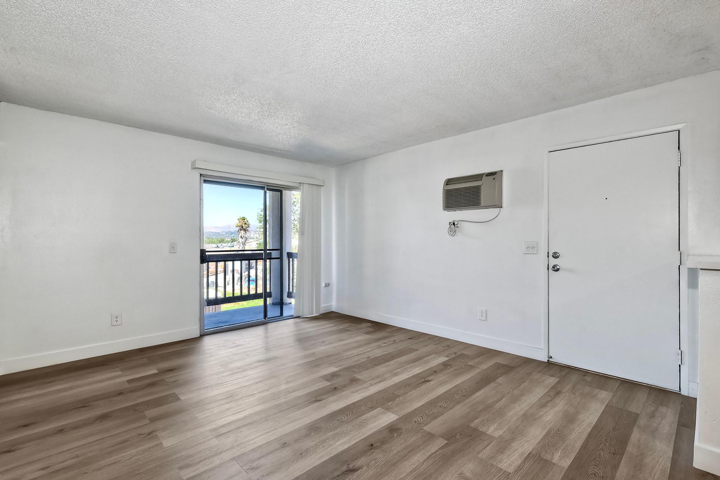 Bright, empty living room featuring light-colored walls and laminate flooring. A door leads to a small balcony with a view. An air conditioning unit is mounted on the wall, and there is a door on the right side of the image. Natural light fills the space.