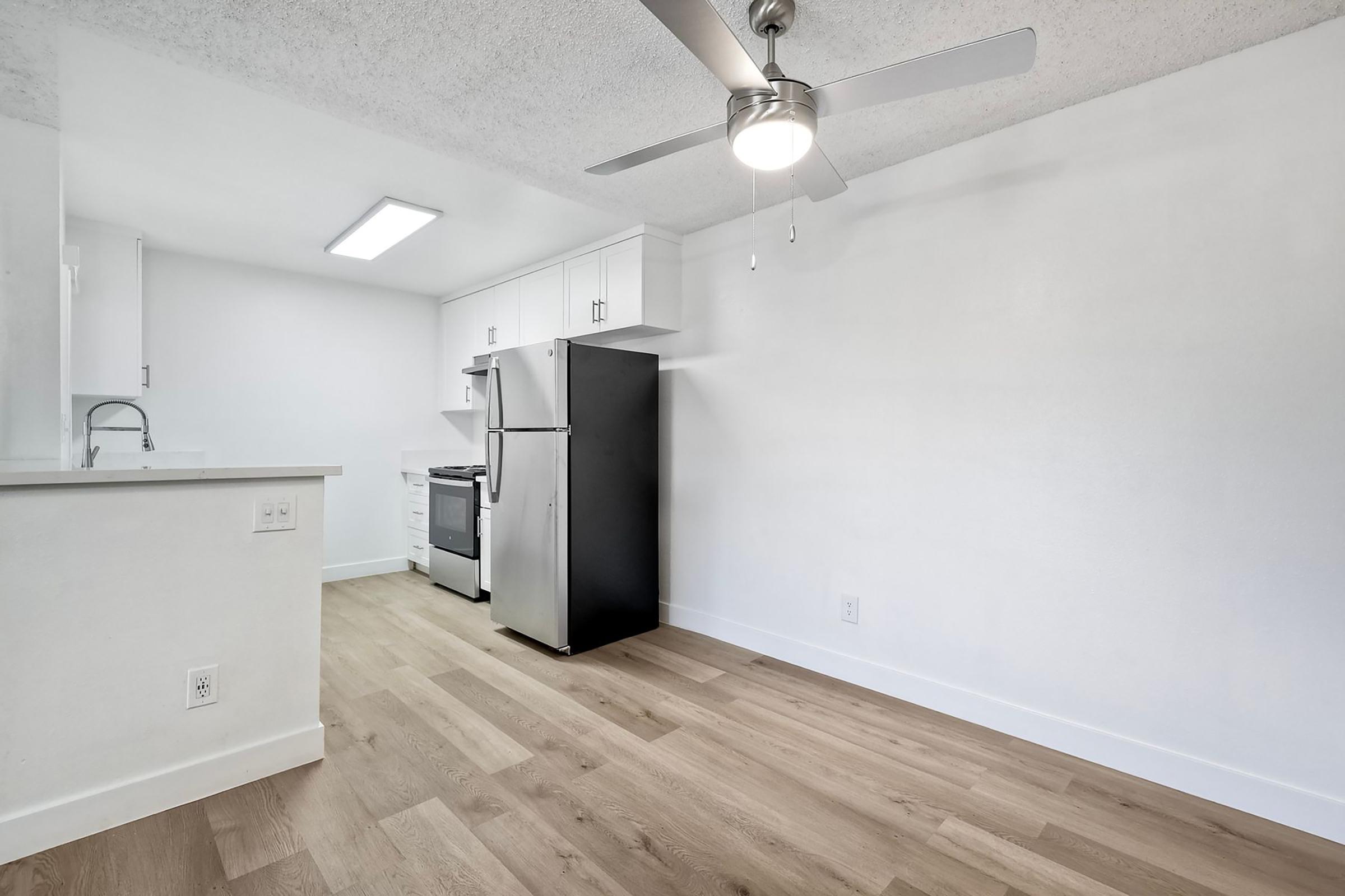 A modern kitchen interior with light wood flooring, featuring a stainless steel refrigerator and stove, white cabinetry, and a ceiling fan. The room is bright and spacious, with a doorway leading to another area.