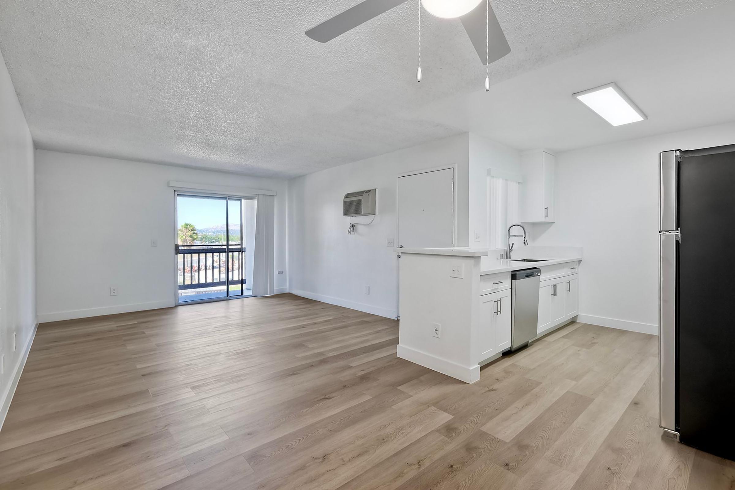 Bright, empty living space with wooden floors, white walls, and a ceiling fan. The kitchen area features modern appliances, including a stainless steel refrigerator and sink. A sliding glass door leads to a balcony with an outdoor view. Natural light fills the room, creating an open and airy atmosphere.