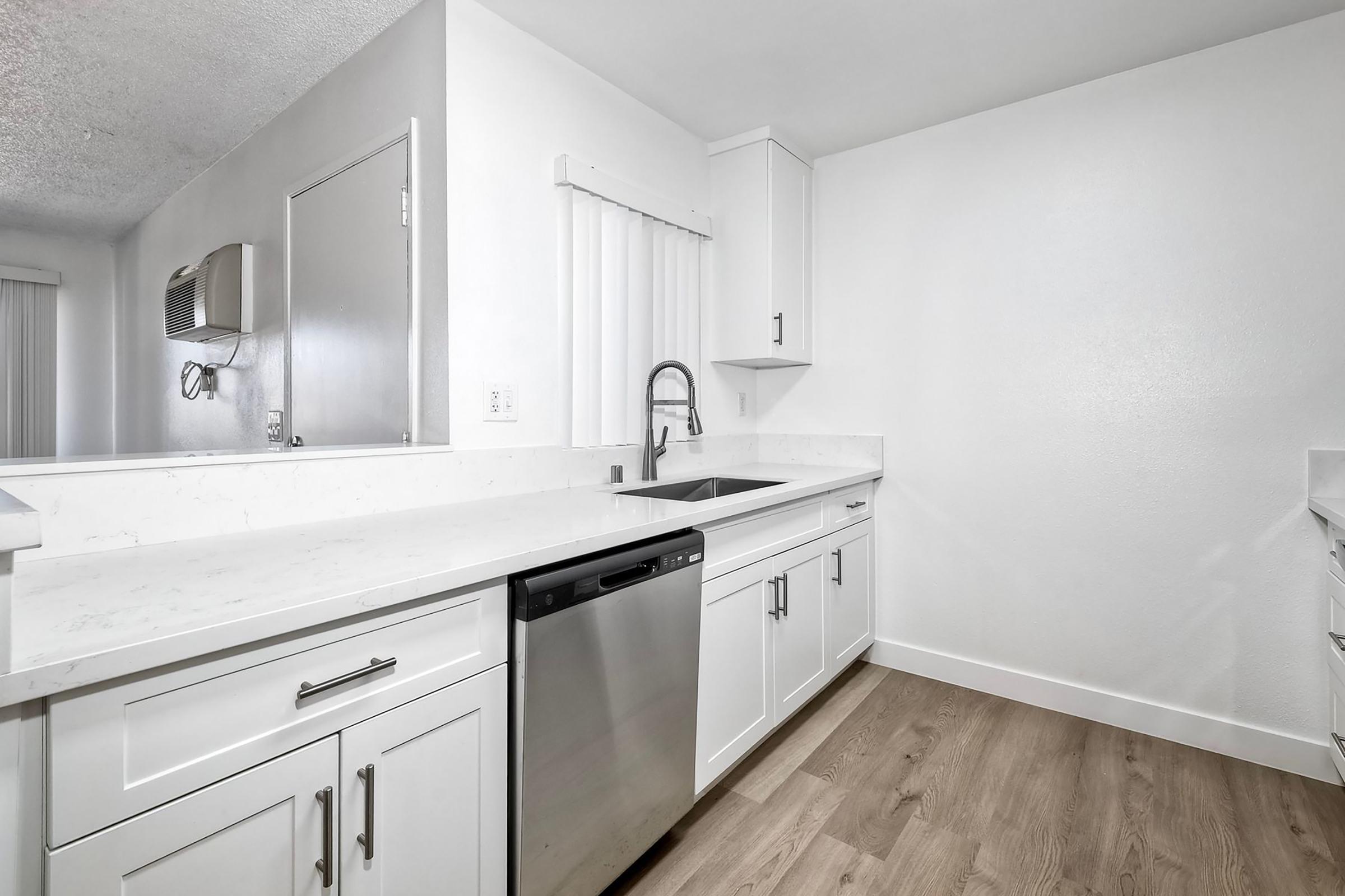 Modern kitchen featuring white cabinetry, a stainless steel dishwasher, and a sleek sink. The design includes a clean countertop with minimal décor, conveying a bright and spacious feel. The walls are painted white, and the floor is made of light wood, enhancing the overall contemporary aesthetic.
