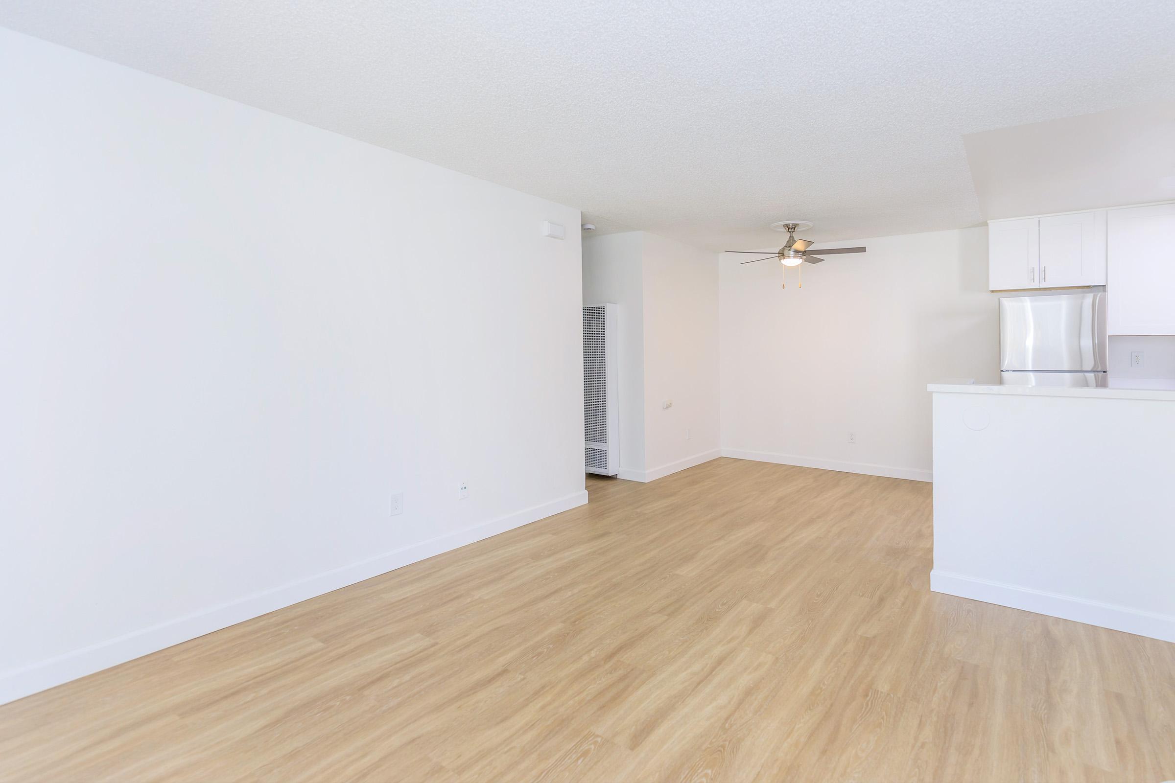 A bright, modern living space featuring light wood flooring and white walls. The room includes a ceiling fan and an open layout leading to a kitchen area with white cabinetry and stainless steel appliances. A doorway in the background suggests access to another room or hallway.