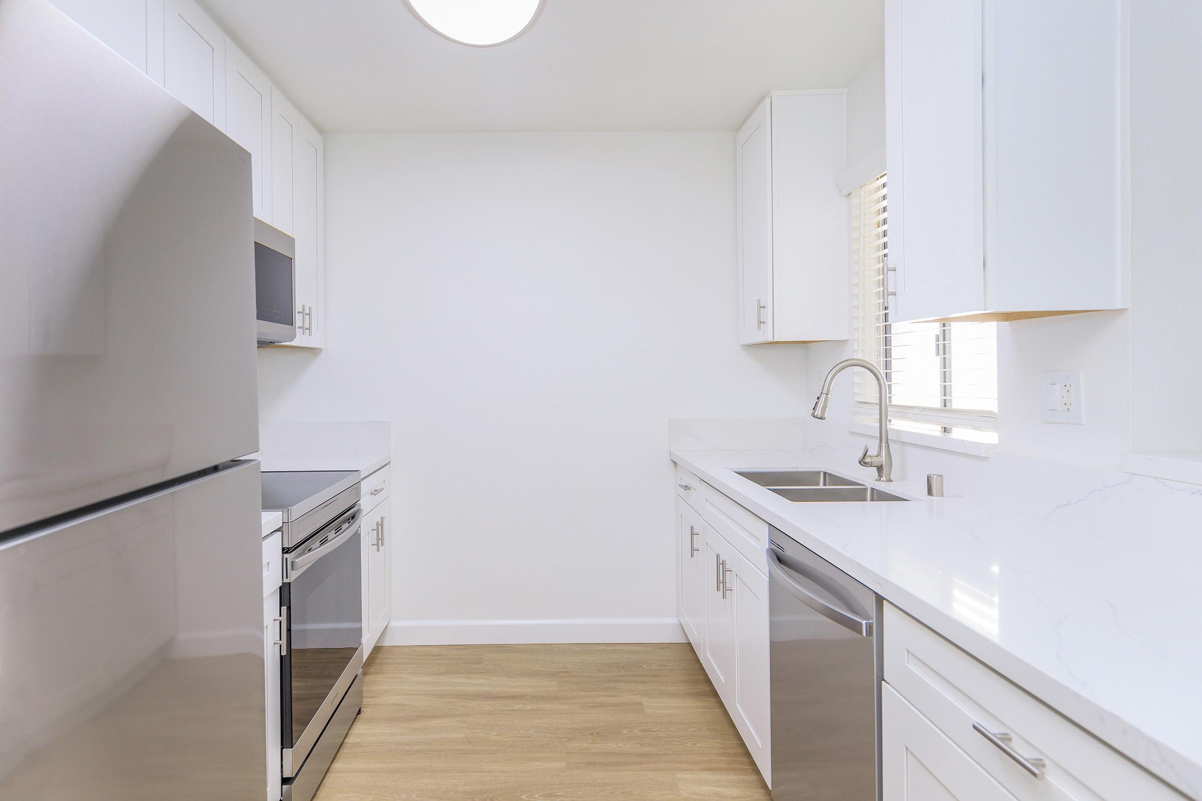 Modern kitchen featuring sleek white cabinetry, stainless steel appliances including a refrigerator, oven, and dishwasher. The countertops are made of white marble, and there's a window allowing natural light. The space has a clean, minimalist design with light-colored wooden flooring.