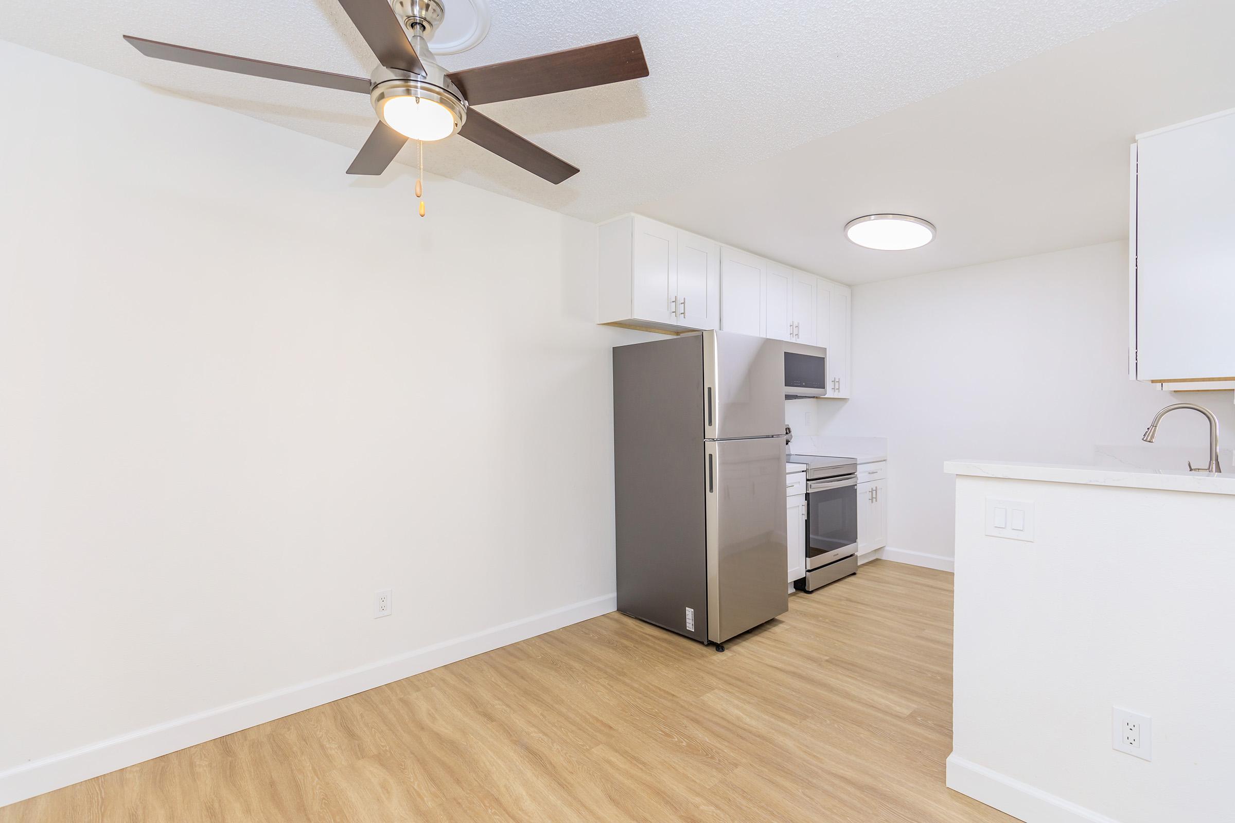 Modern kitchen with a stainless steel refrigerator and stove, white cabinets, and a light wooden floor. A ceiling fan is visible, and the walls are painted white, giving a bright and spacious feel to the room.