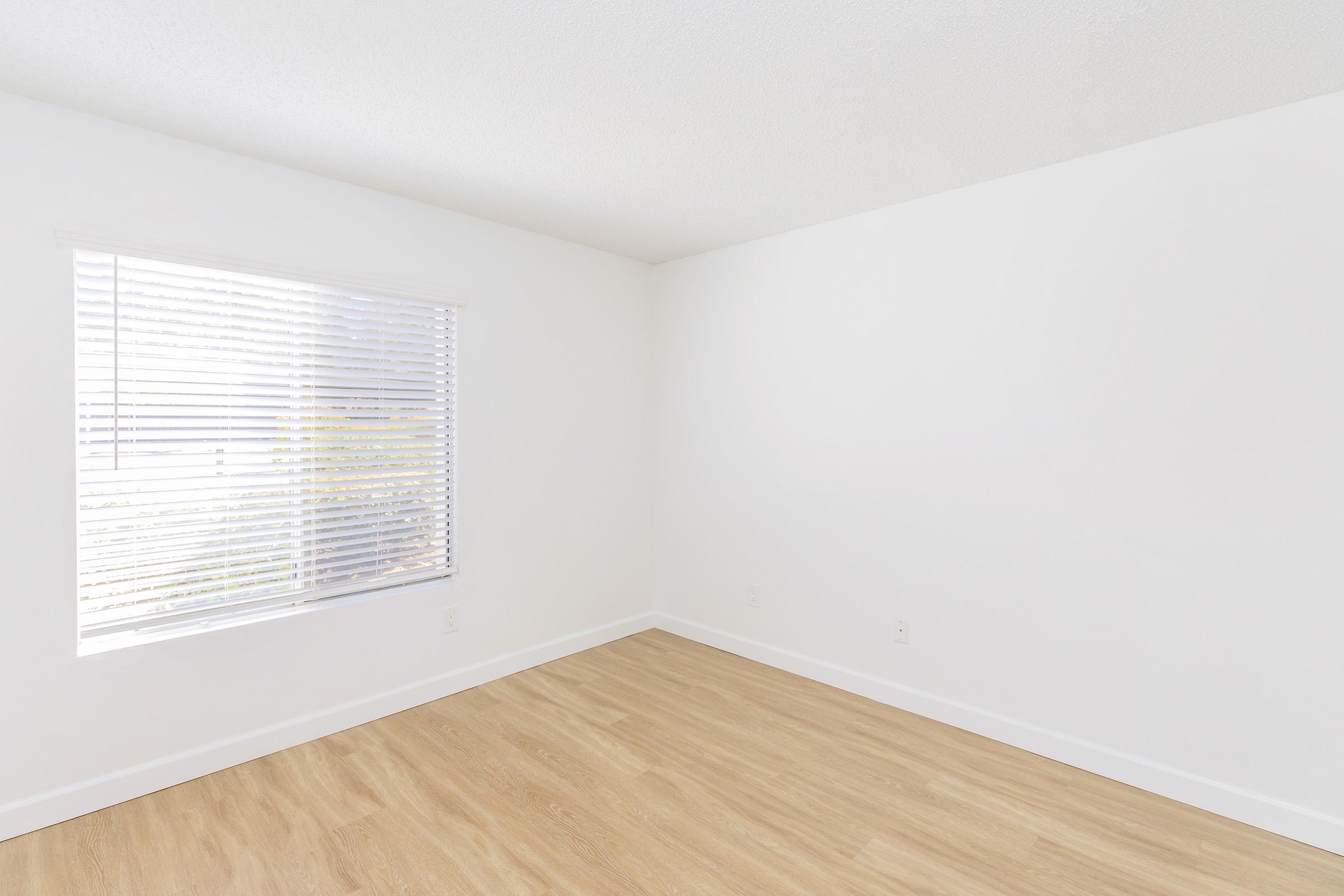 A bright, empty room with white walls and light wooden flooring. It features a large window with white blinds allowing natural light to enter. The space has a clean and minimalist aesthetic, suitable for various uses.