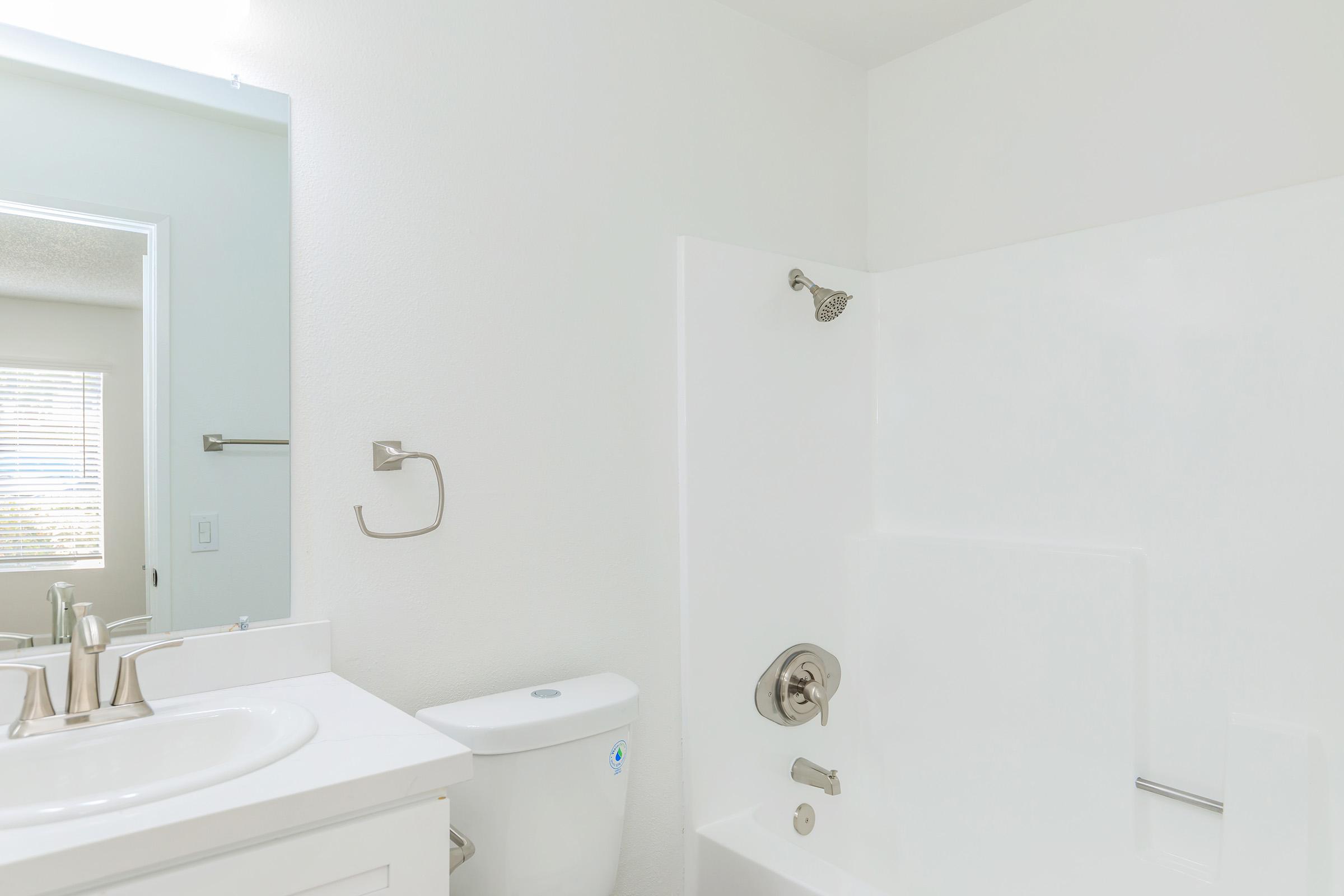 A clean and modern bathroom featuring a white bathtub with a shower, a sink with two faucets, and a toilet. The walls are painted white, and there is natural light coming from a window, reflecting a minimalist design.