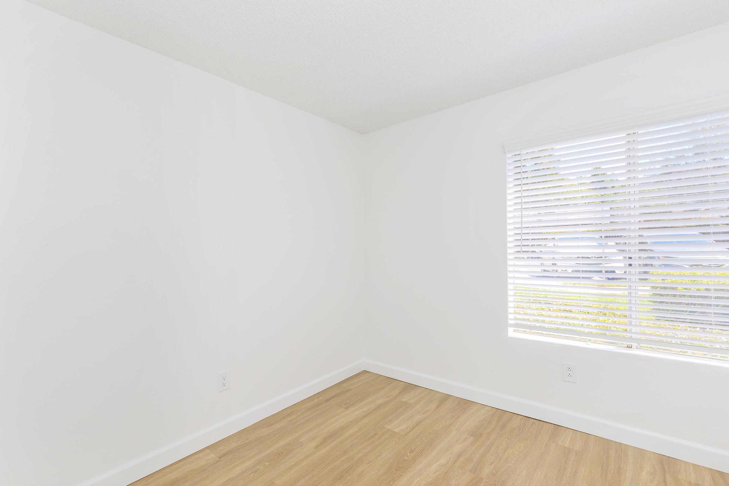 A bright, empty room with white walls and light brown wooden flooring. A window with blinds allows natural light to enter, illuminating the space. The room has a clean and minimalist aesthetic, perfect for various uses or personalization.