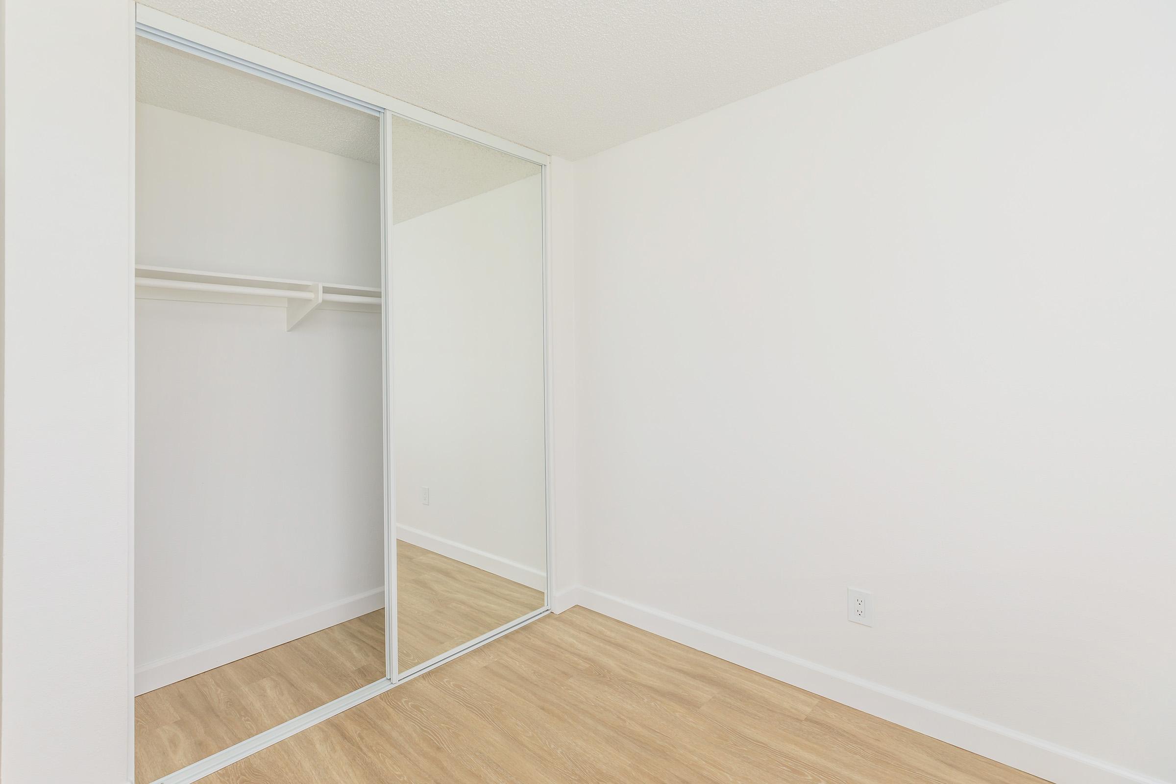 A bright, empty room with light-colored walls and a wooden floor. On one side, there is a sliding mirrored closet with a simple shelf inside. The space has a clean and minimalist appearance, with ample natural light enhancing the openness of the area.