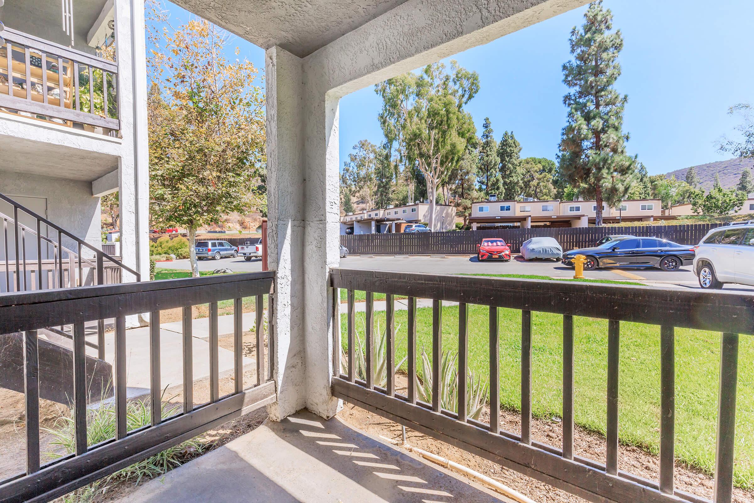 View from a balcony or porch, featuring a concrete railing and a grassy area below. In the background, there are parked cars along a tree-lined street. Clear blue sky and greenery are visible, creating a pleasant outdoor scene.