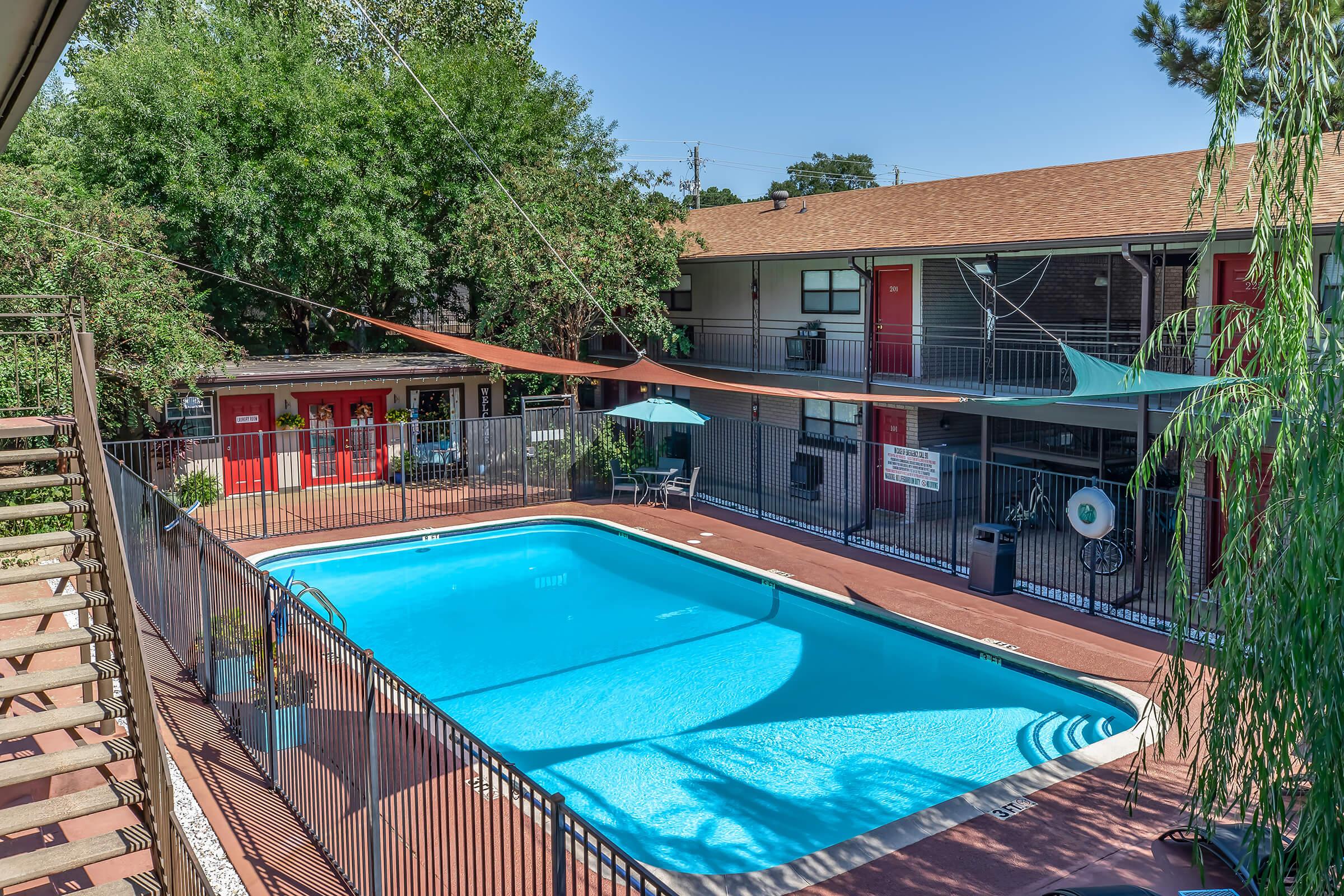 A sunny courtyard featuring a clear blue swimming pool surrounded by a black fence. Nearby are lounge chairs and umbrellas, with an apartment building in the background that has red doors and green accents. Lush trees provide shade, creating a relaxing atmosphere.