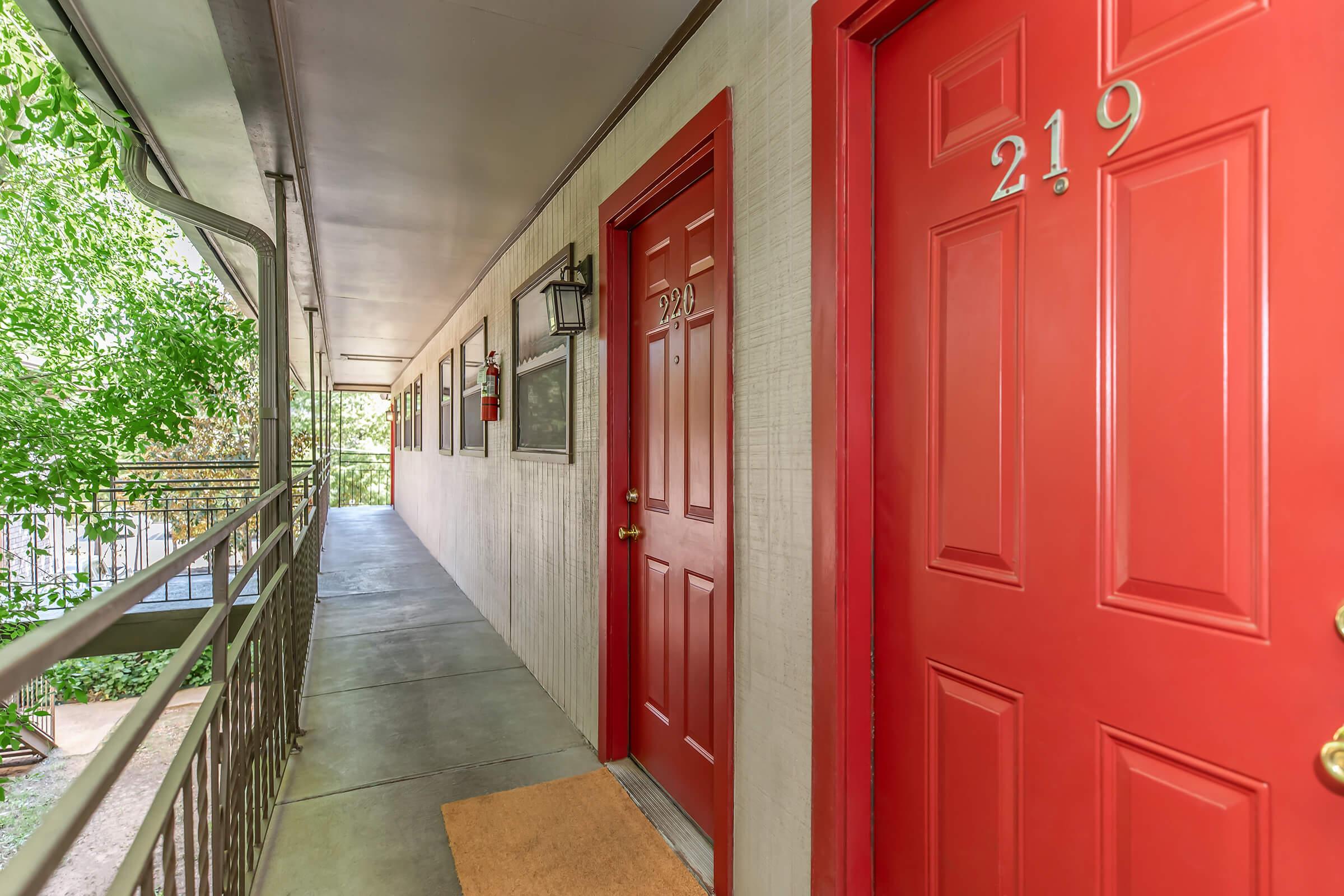 A view of a corridor in an apartment building featuring two red doors, numbered 219 and 214. The walkway is lined with greenery and has a light fixture on the wall. The corridor has a simple, clean design with a concrete floor and a doormat at the entrance.