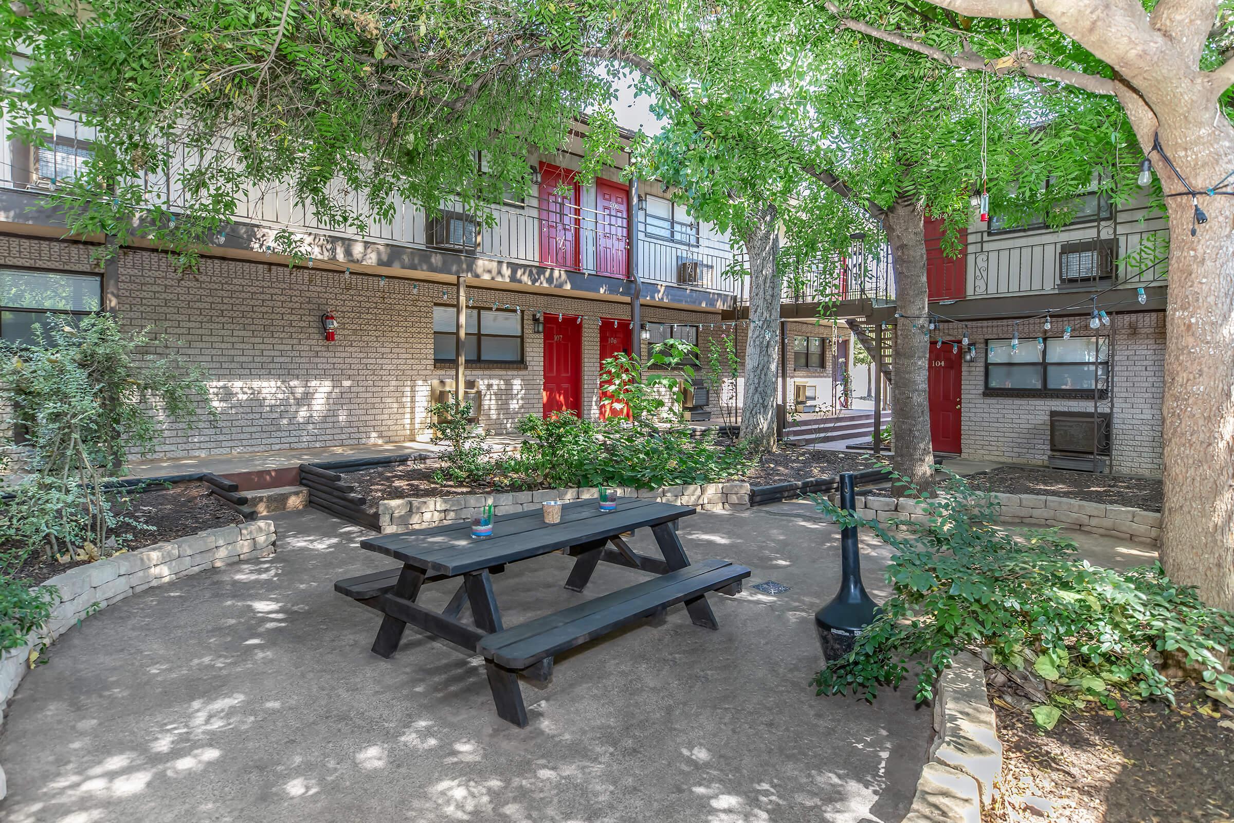 A tranquil courtyard featuring a wooden picnic table surrounded by greenery. Two stories of apartment buildings are visible in the background, with some units having red doors. Soft lighting hangs from trees, adding a cozy atmosphere to the space, perfect for outdoor gatherings.