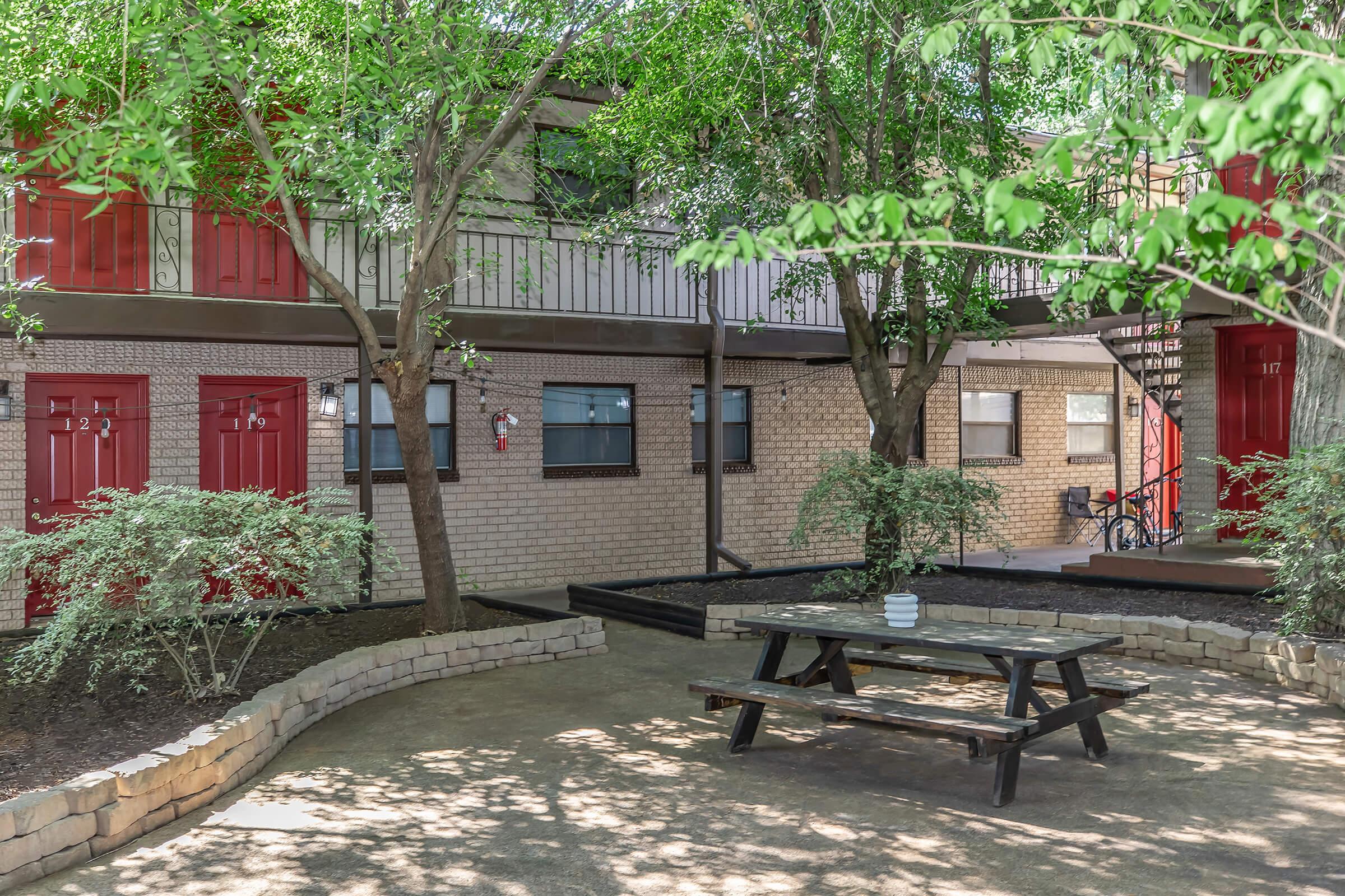 A tranquil outdoor area featuring a picnic table surrounded by greenery, with two-story buildings in the background. The buildings have red doors and windows, and a staircase is visible on the right. The setting is shaded by trees, creating a peaceful atmosphere.