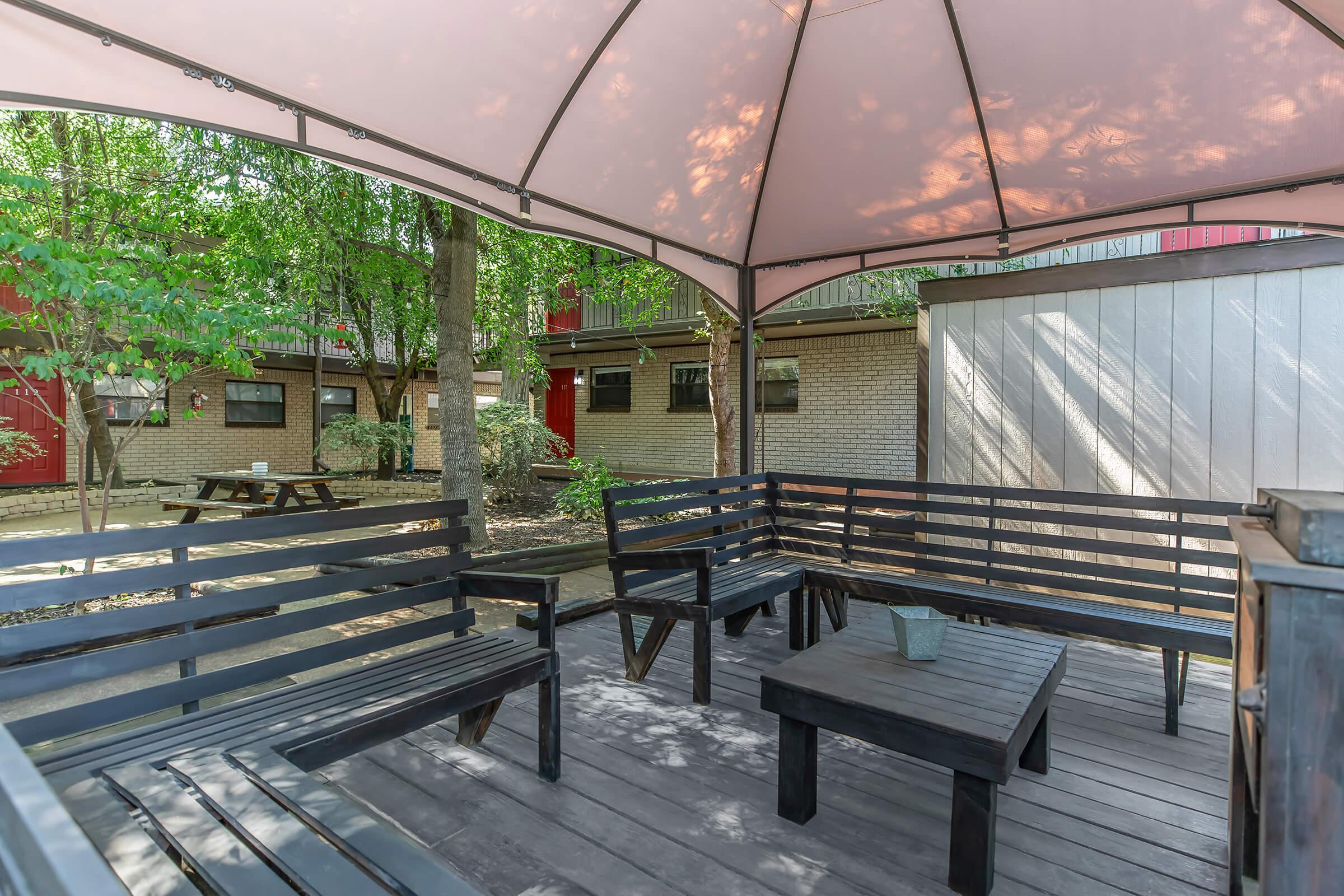 A shaded outdoor sitting area with black wooden benches and a table, surrounded by greenery. The gazebo structure provides shelter, while nearby buildings and picnic tables are visible in the background. Natural light filters through trees, creating a tranquil atmosphere.