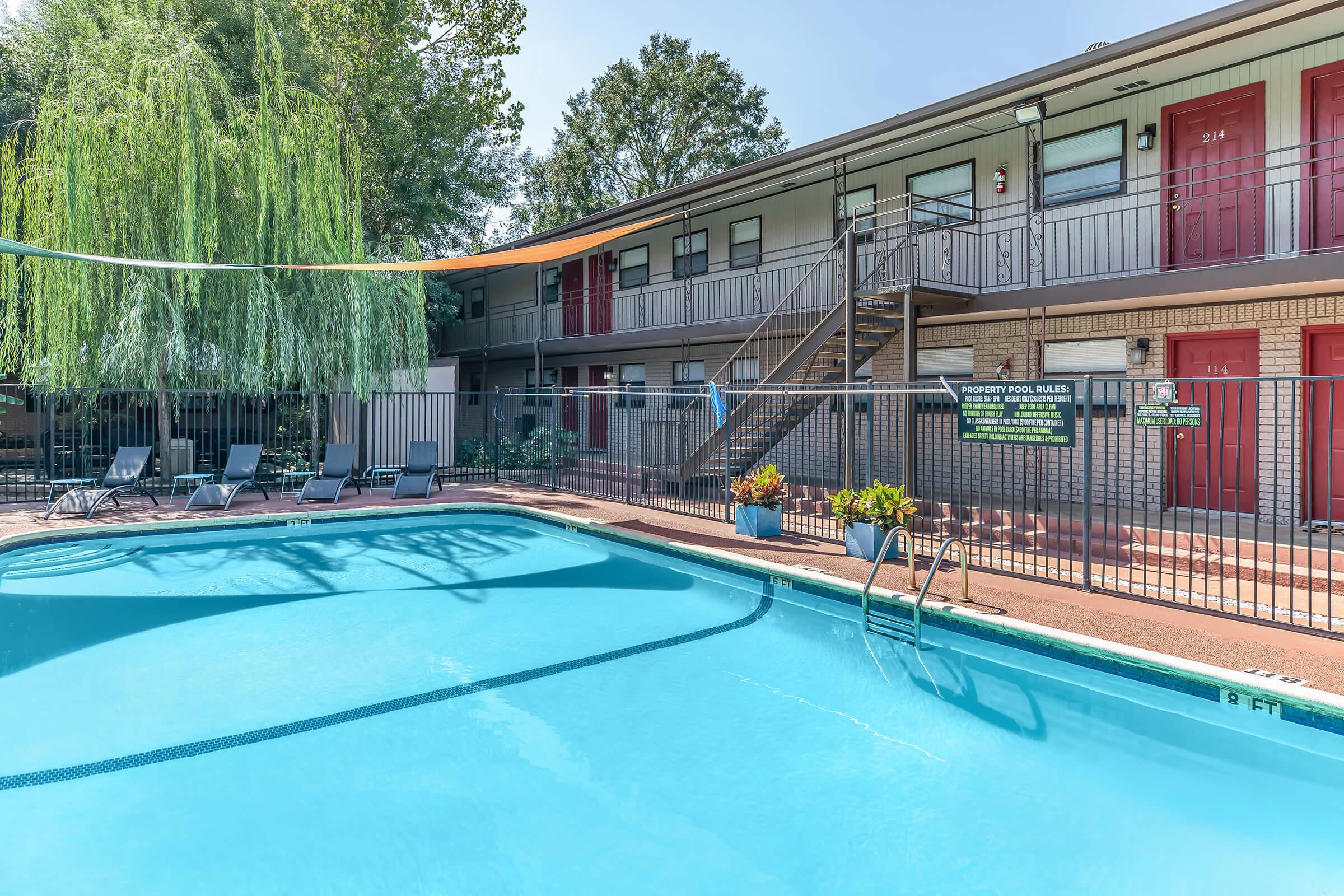 A clear blue swimming pool surrounded by lounge chairs is situated in front of a two-story building with red doors. Lush green trees provide shade, and a sign detailing property pool rules is visible near the pool area. The scene conveys a relaxed, inviting atmosphere.