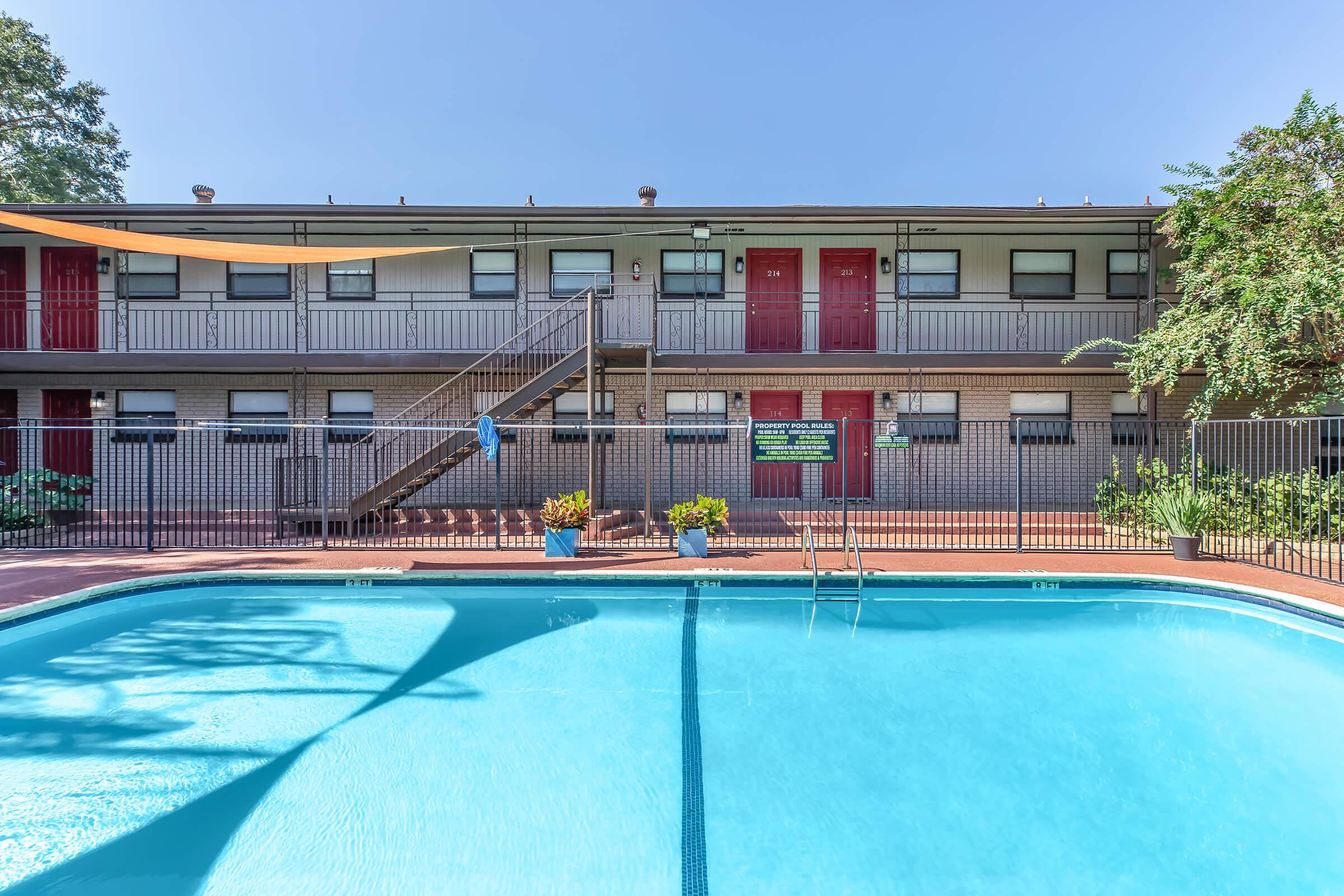 A clear blue swimming pool in the foreground with a surrounding fence. Behind the pool, there are two levels of an apartment building featuring red doors and a staircase. The area is well-lit with green plants and a sunny sky above. Simple outdoor decor enhances the inviting atmosphere.