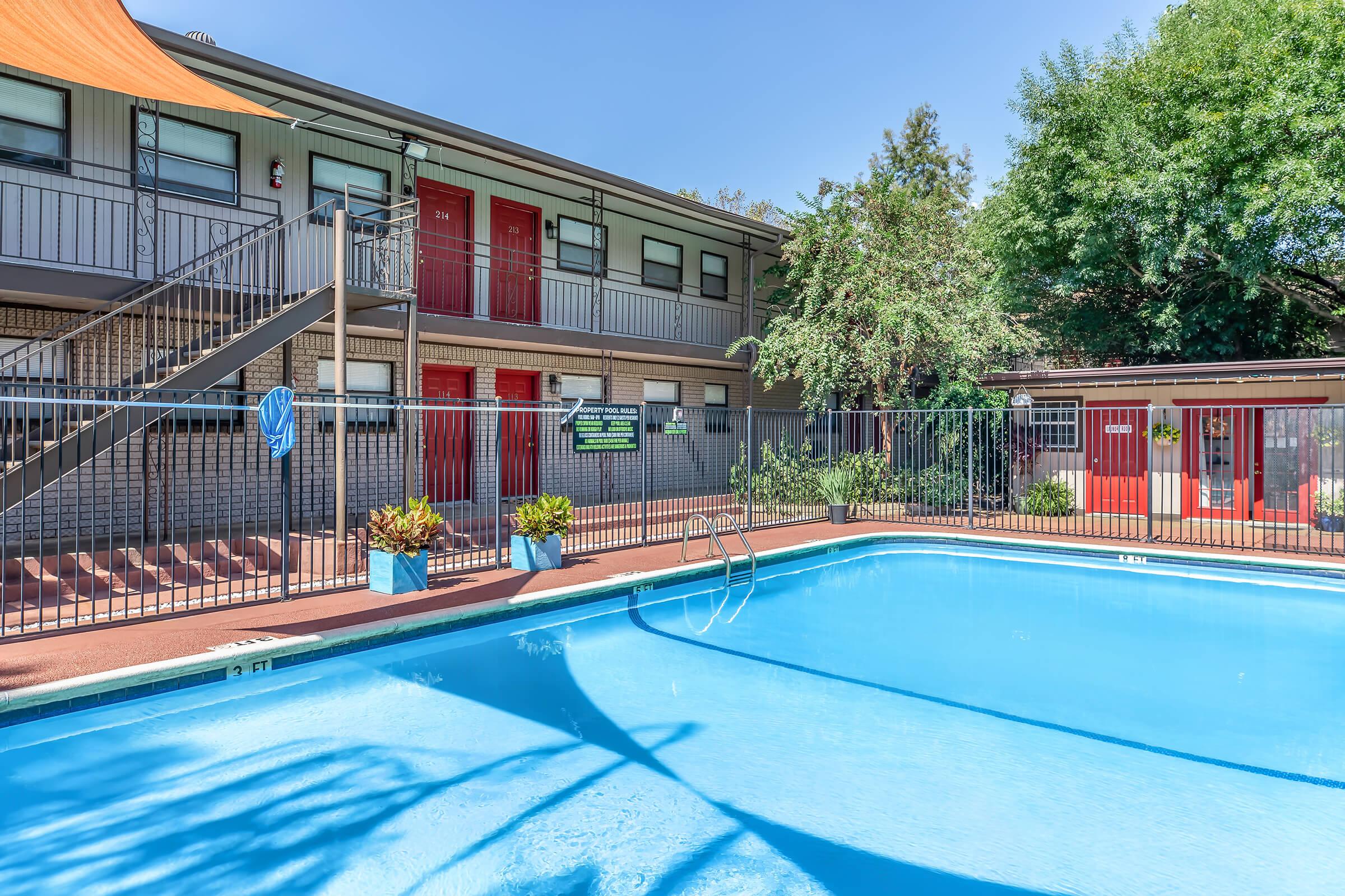 A swimming pool surrounded by a fenced area, with a staircase leading to a two-story building featuring red doors. Lush greenery is visible around the pool area, and there are potted plants near the edge of the pool. The scene is set on a clear, sunny day.