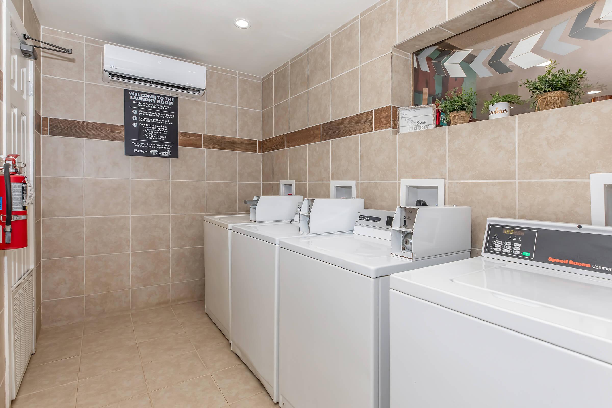 A clean, modern laundry room featuring multiple white washing machines and dryers. The wall is tiled with beige tiles, and there's a large mirror above the appliances. A sign on the wall provides instructions or information about laundry services, and there are decorative plants in the corner.