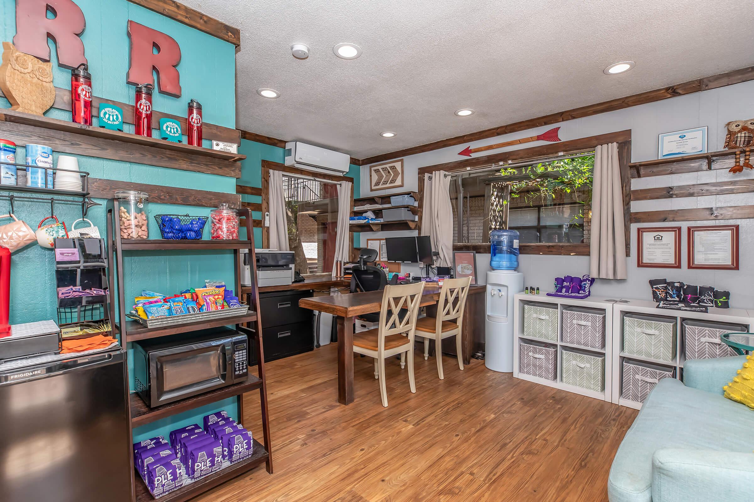 A cozy interior of a small office or break room featuring shelves with snacks and drinks, a microwave, and a water cooler. Two chairs are positioned at a desk with a computer, and decorative elements include wooden accents and colorful wall decor. Bright natural light enters through a window.