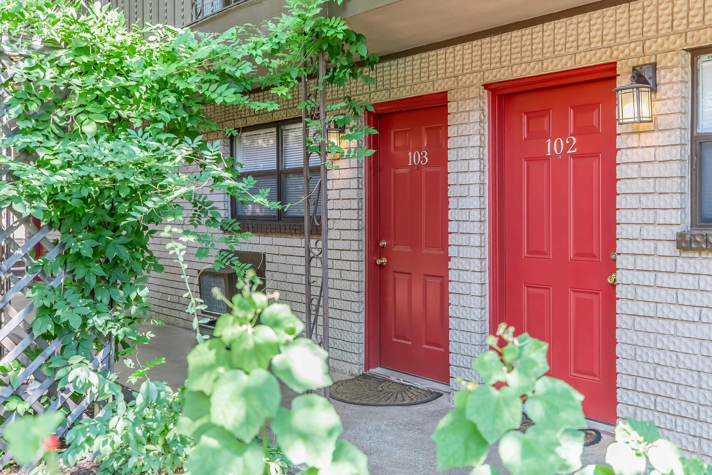 Two red doors numbered 102 and 103 on a textured beige exterior wall of a building. Green foliage and plants are visible in the foreground, adding a touch of nature. The entrance is well-lit with an outdoor light beside each door. The scene suggests a welcoming atmosphere.